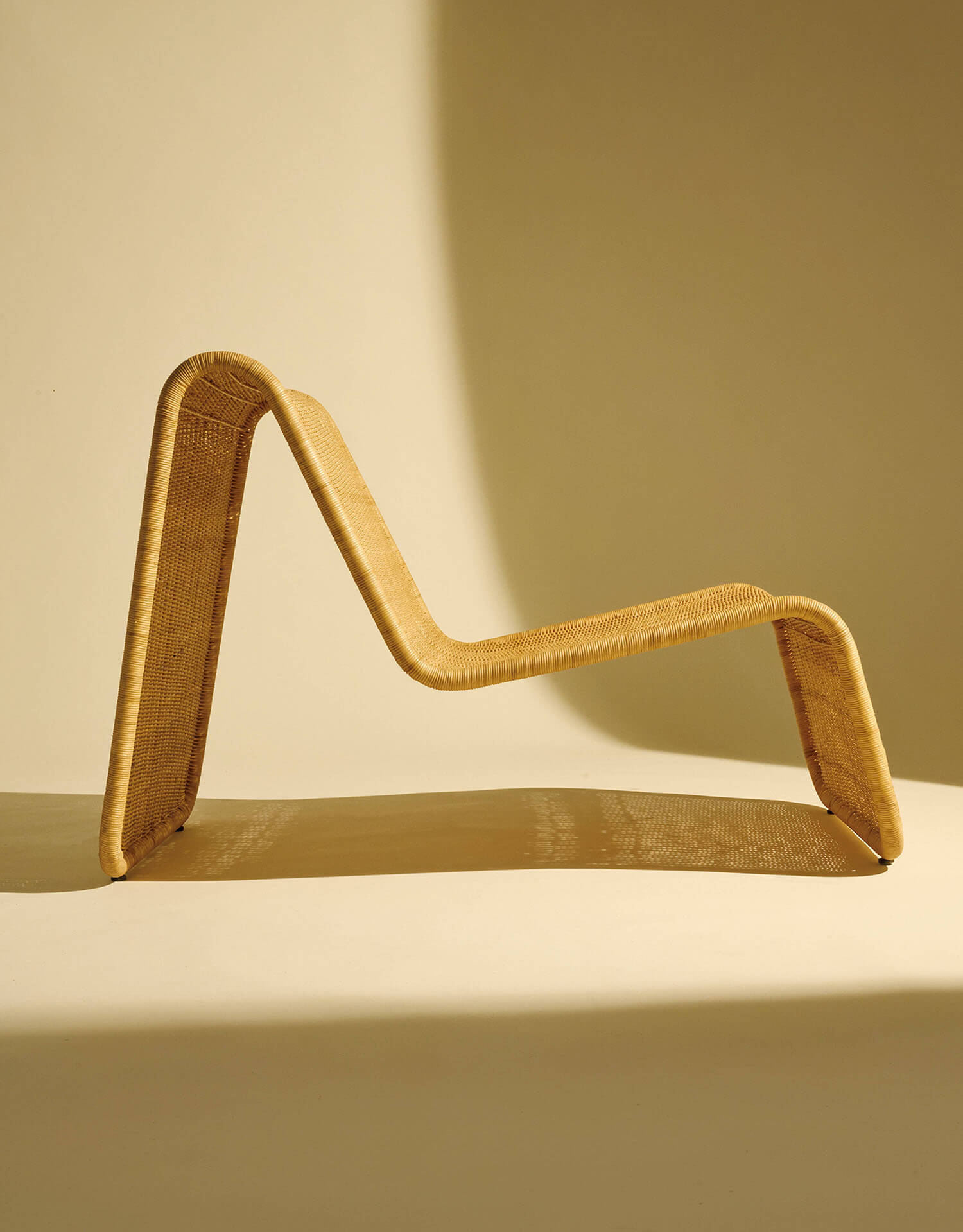 A modern, sculptural lounge chair made of woven rattan, with angular, continuous curves, set against a beige background with soft shadows.