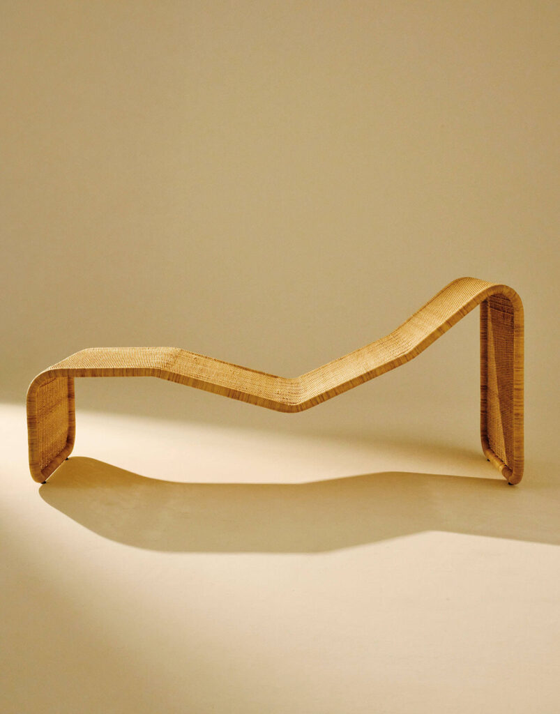A sculptural, minimalist wooden chaise lounge with a zigzag form is displayed against a plain beige background.