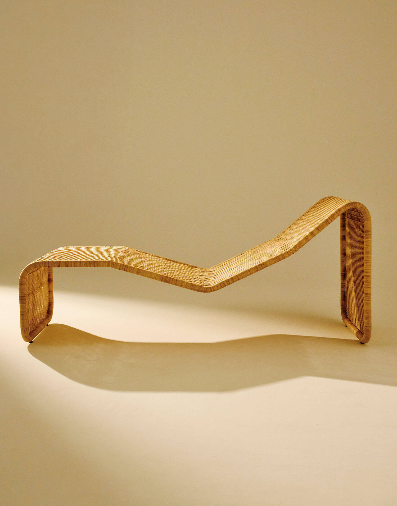 A sculptural, minimalist wooden chaise lounge with a zigzag form is displayed against a plain beige background.