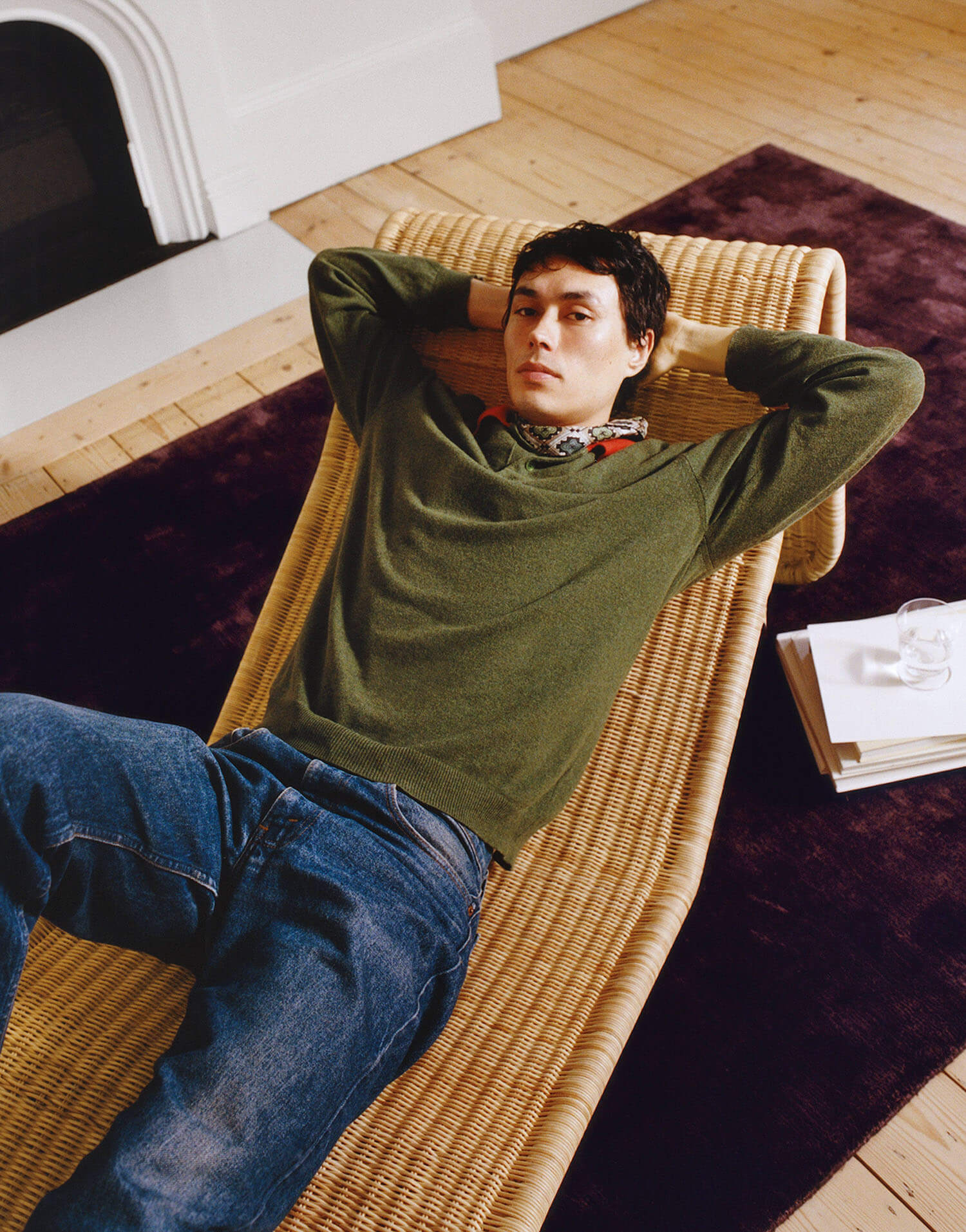 A person in a green sweater and blue jeans lies on a woven chaise lounge with hands behind their head, next to a purple rug and a small table with books and a glass of water.