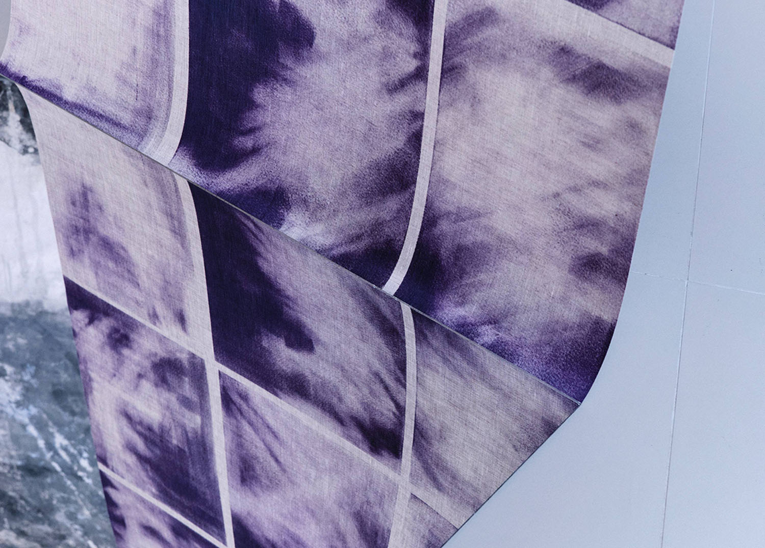 A close-up of purple and white fabric with a grid pattern, draped over a surface. The fabric features tie-dye-like, cloudy texture within each square.
