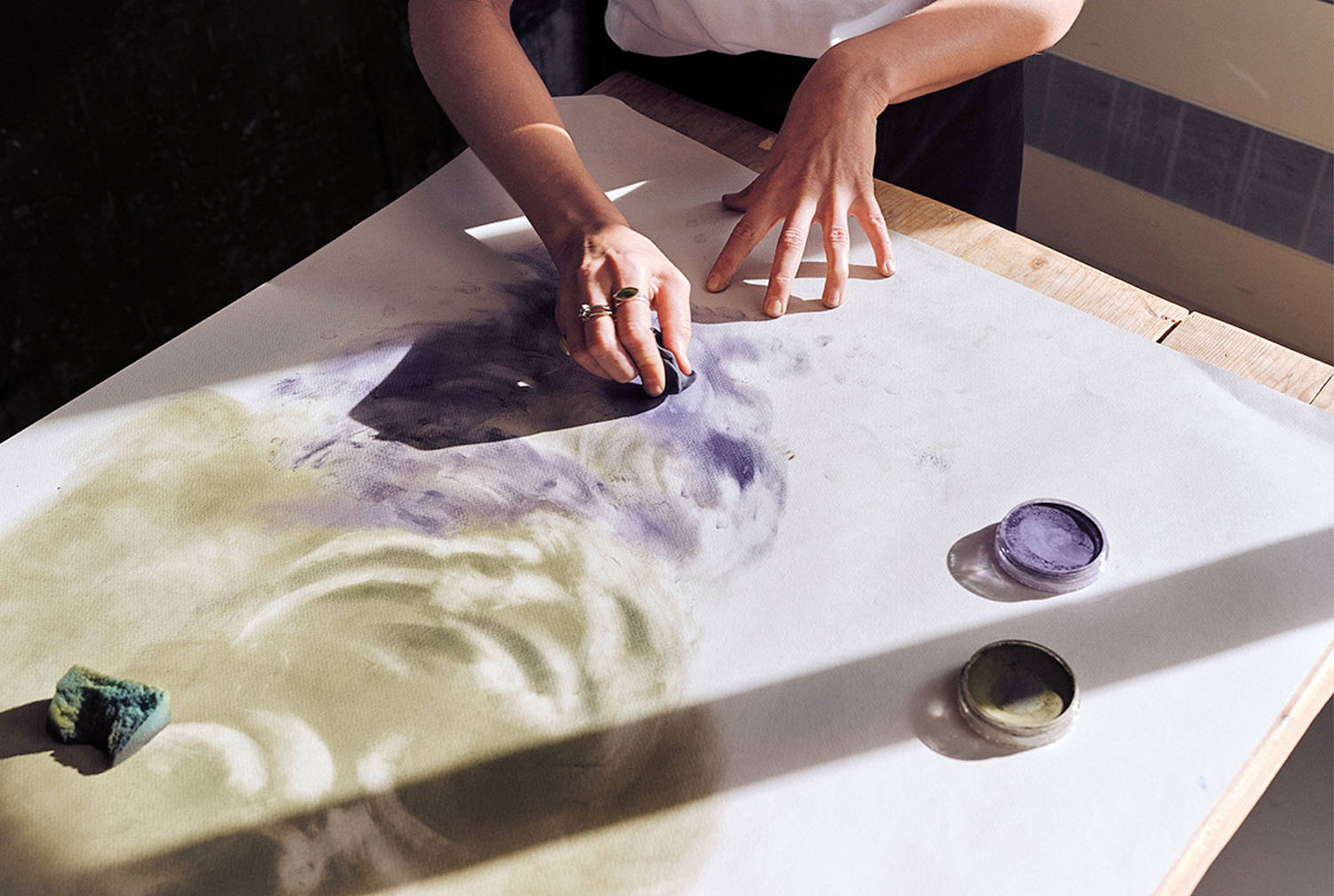 A person uses their hands to blend purple and green pastels on a large sheet of paper, with pastel containers and a sponge nearby on a wooden table.