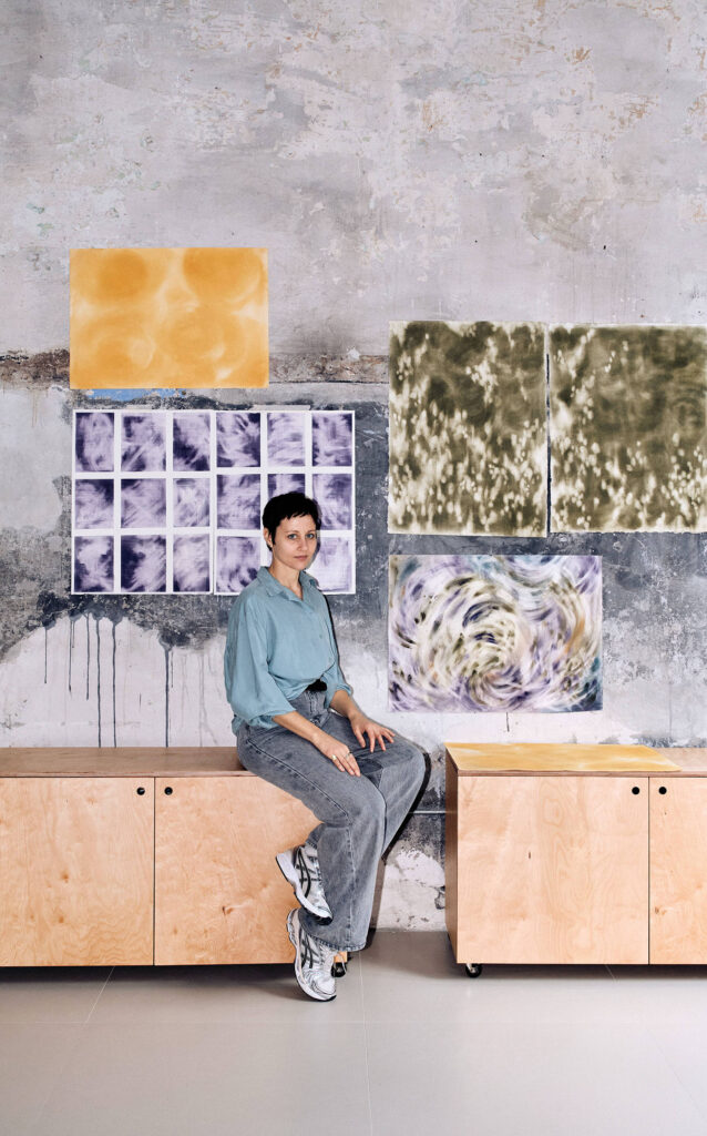 A person in a blue shirt and gray pants sits on a wooden cabinet in front of a wall with abstract art pieces.