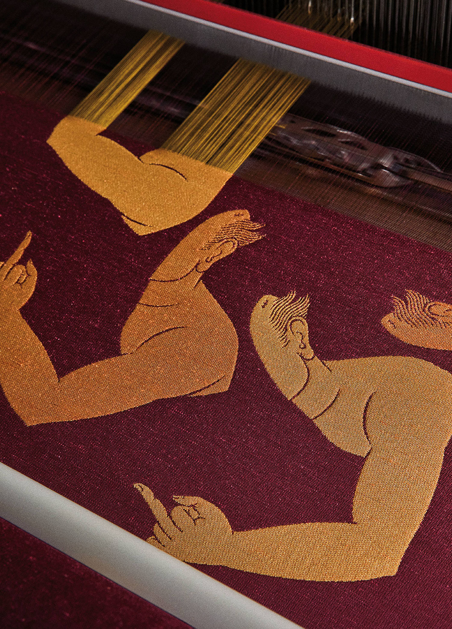 A maroon textile with gold woven figures of stylized arms and faces, shown in profile, under the threads of a weaving loom.