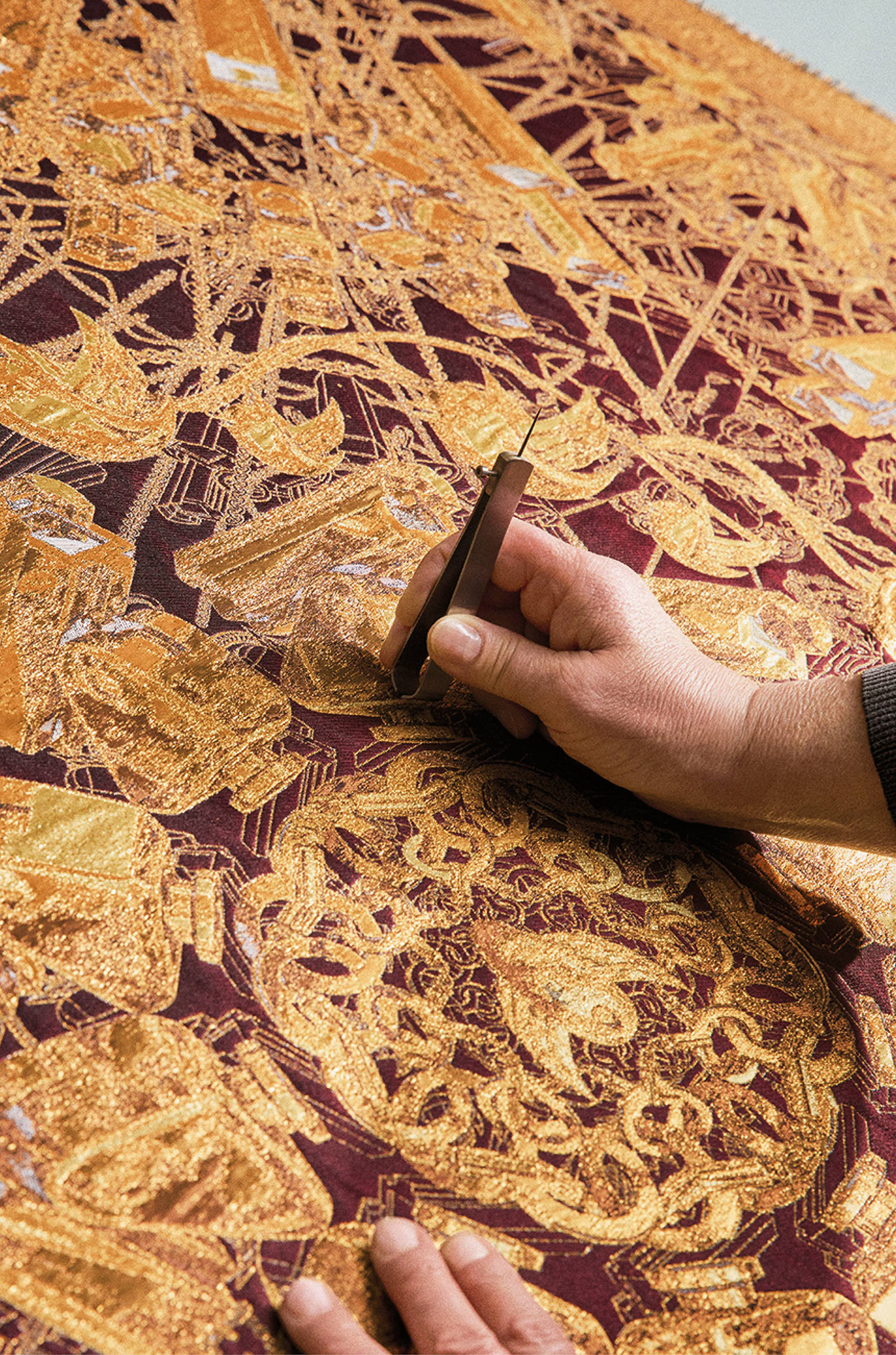 A person uses a tool to work on intricate gold embroidery on a rich maroon fabric.
