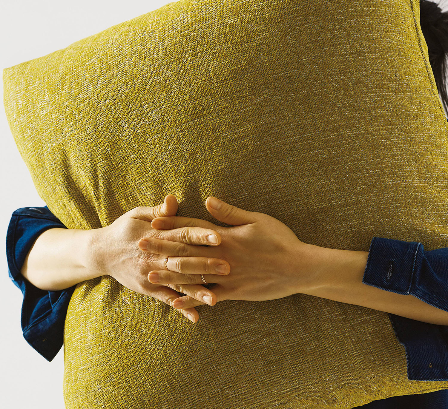 A person wearing a blue shirt holds a large yellow pillow, with both hands clasped around it against a plain light background.