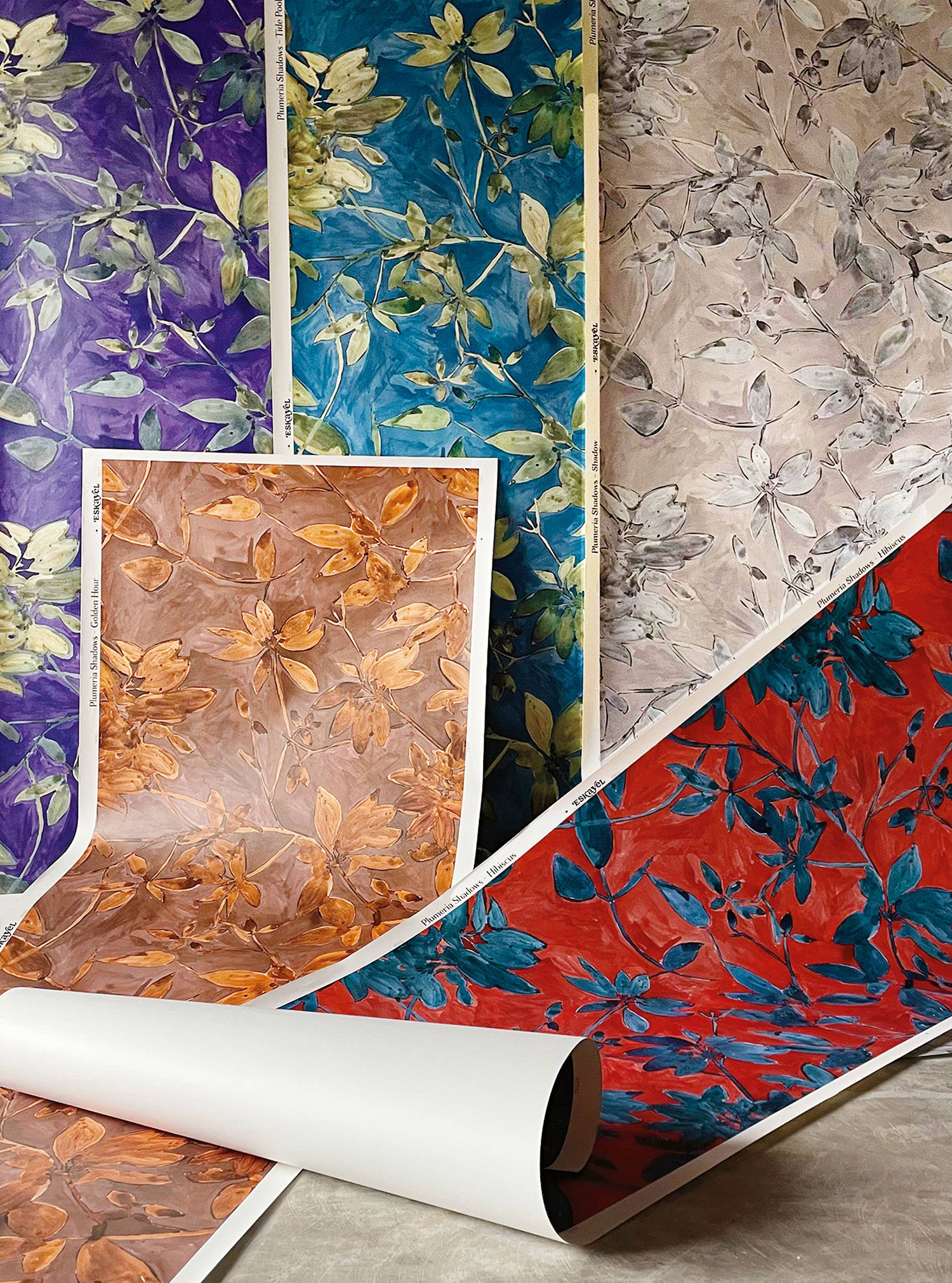 Five rolls of floral wallpaper in purple, teal, beige, orange, and red, each featuring a botanical leaf and flower pattern, are displayed upright and partially unrolled on a surface.