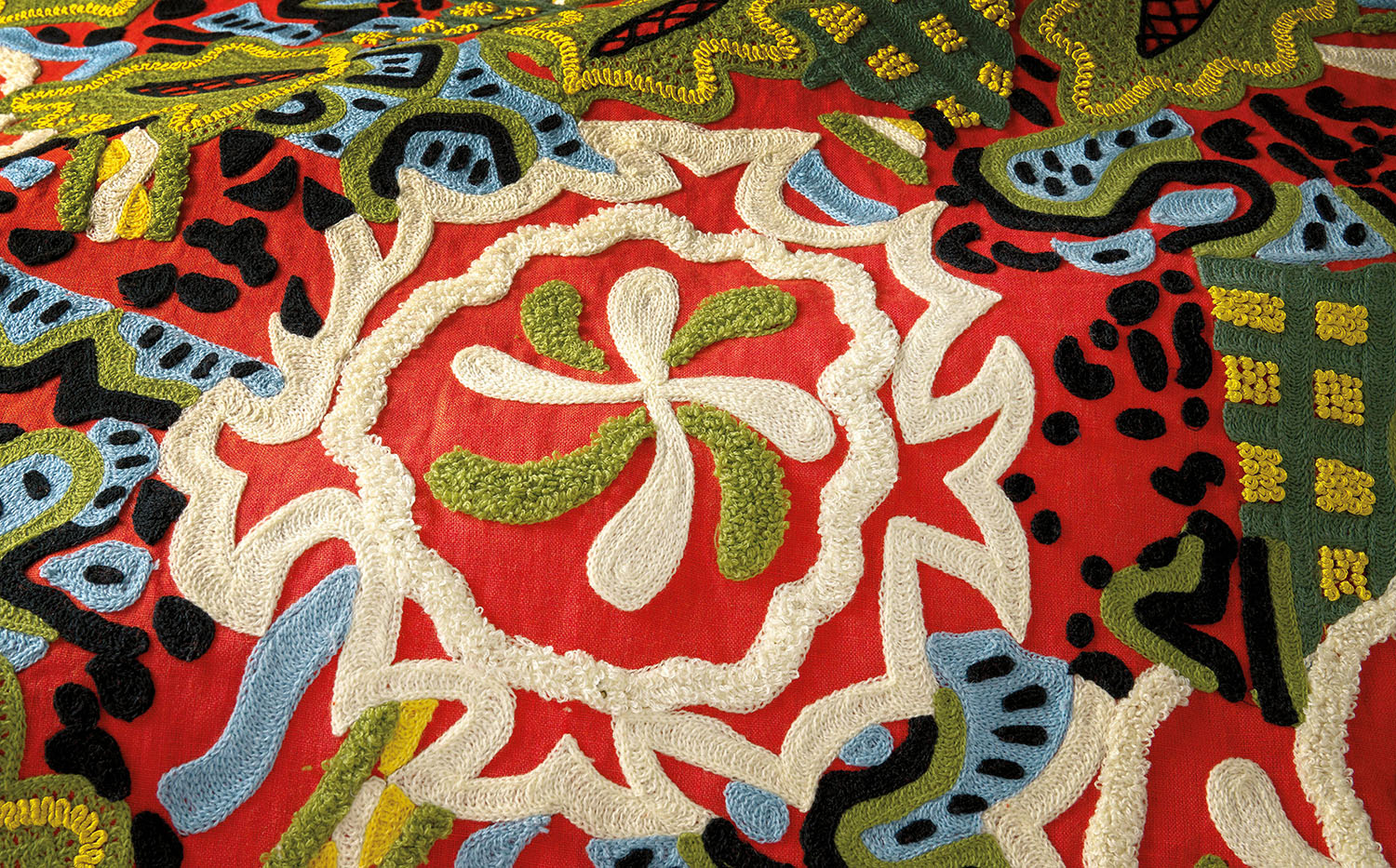 Colorful textile with intricate embroidered patterns in red, white, green, blue, and black, featuring floral and abstract shapes.