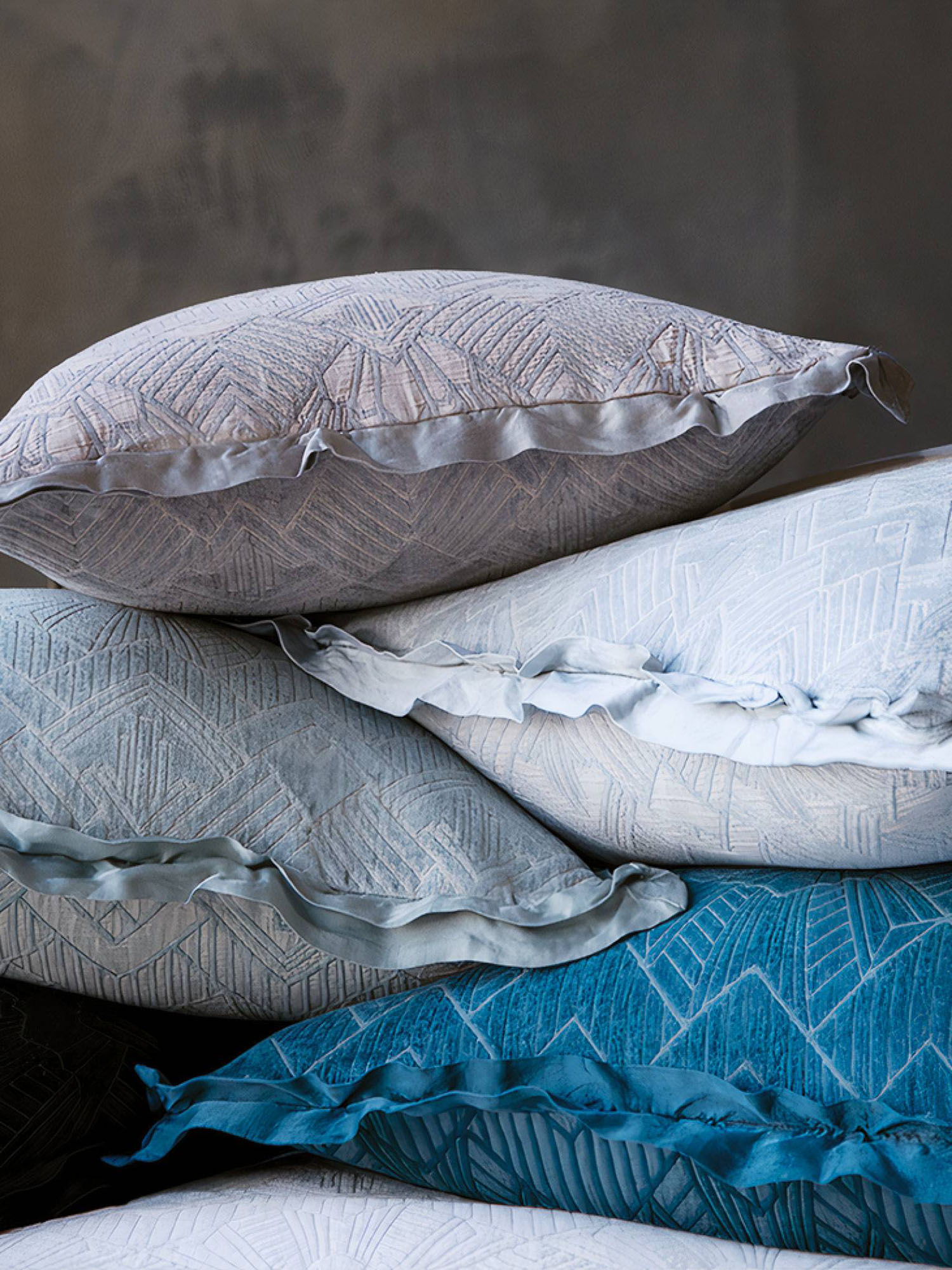 A stack of textured pillows in shades of blue, grey, and beige with subtle geometric patterns and ruffled edges against a dark background.
