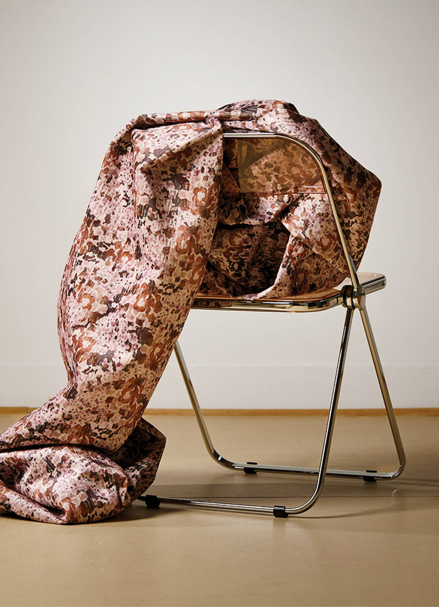 A metal folding chair with a large, patterned pink and brown fabric draped over and around it, set against a plain wall and floor.