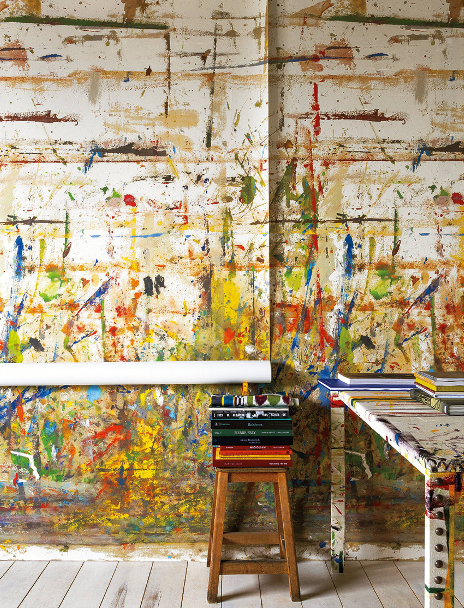 Room with paint-splattered walls, a matching table, and a wooden stool stacked with books; bright colors and abstract patterns dominate the scene.