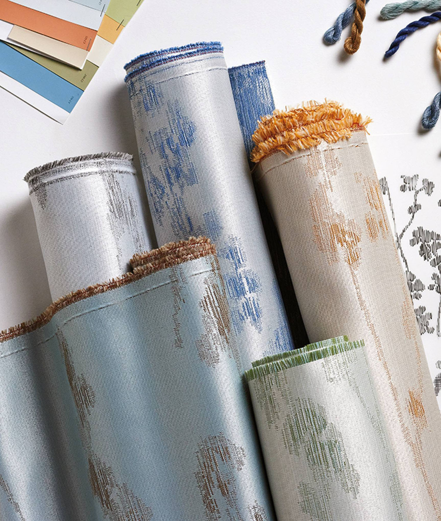 Several rolled-up fabric samples with various patterns and trims are displayed on a table alongside color swatches and embroidery threads.