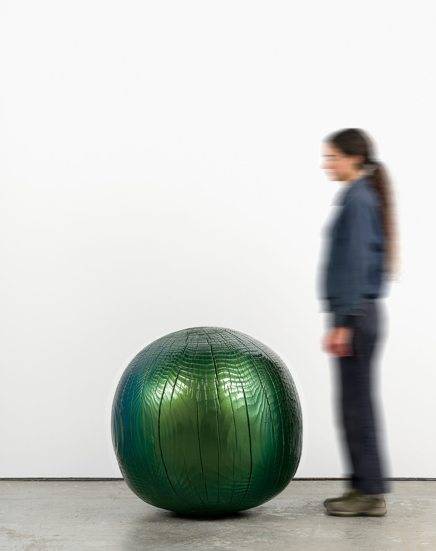 A person standing next to a large, glossy green spherical sculpture with a textured surface, in a minimalist indoor setting.