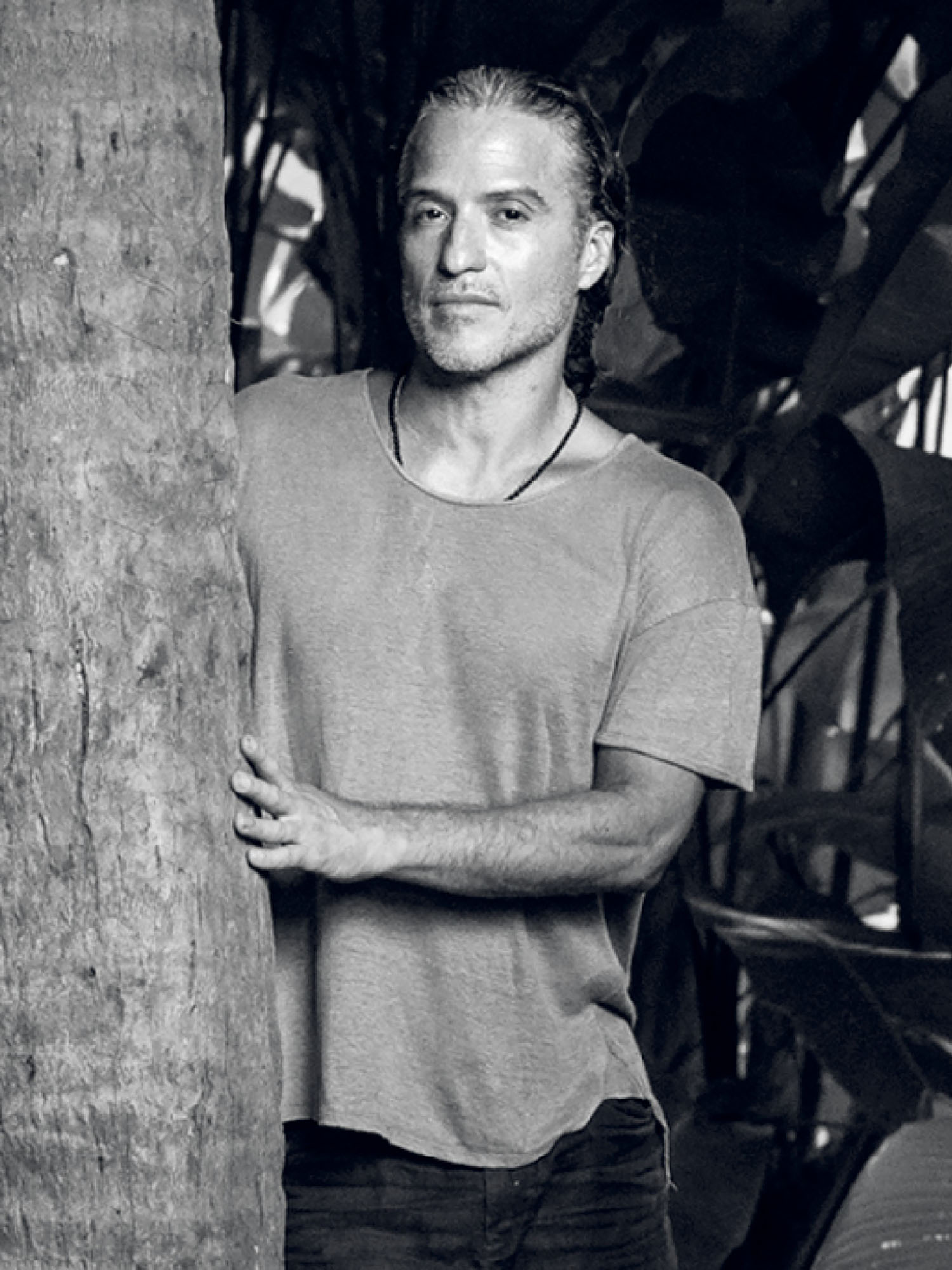 A man with long hair stands outdoors, leaning against a tree, wearing a casual t-shirt and necklace, with large leafy plants in the background.