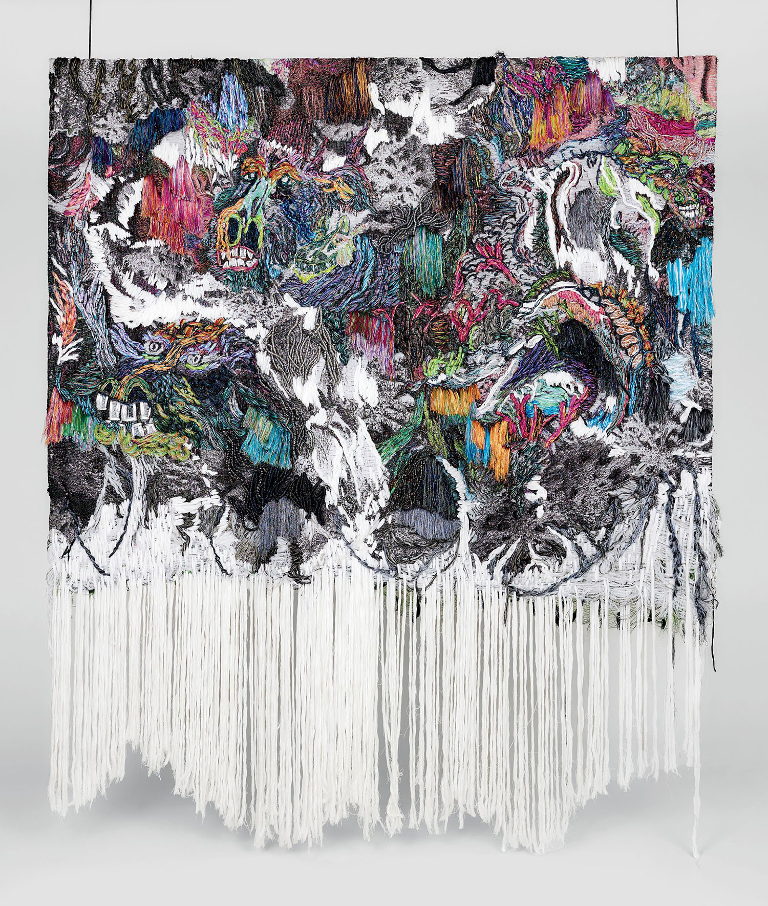 A colorful, abstract textile artwork with dense, intricate embroidery on top and long, white yarn strands hanging loosely from the bottom.