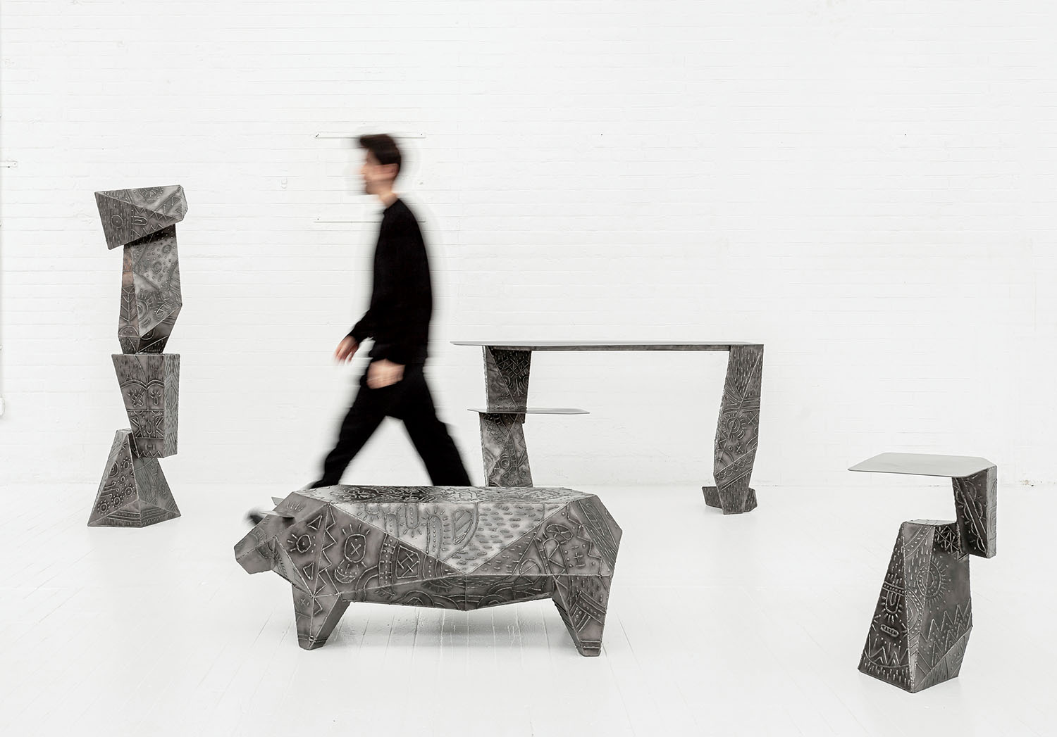 A person in black walks past angular, sculptural metal furniture pieces in a white, minimalist room.