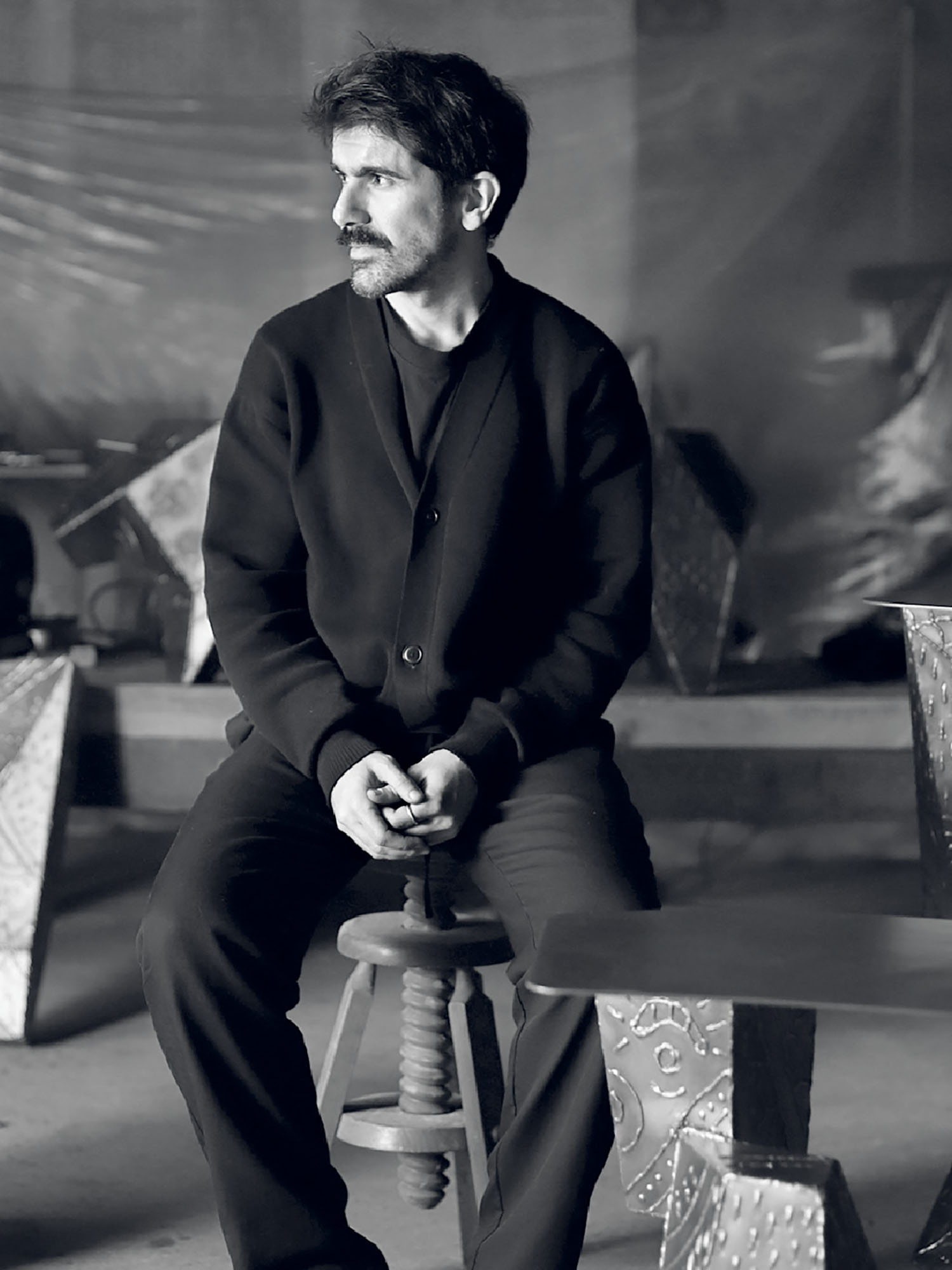 A man with dark hair and a mustache sits on a wooden stool indoors, wearing a dark jacket and pants, with geometric metal tables nearby. The background is dimly lit.