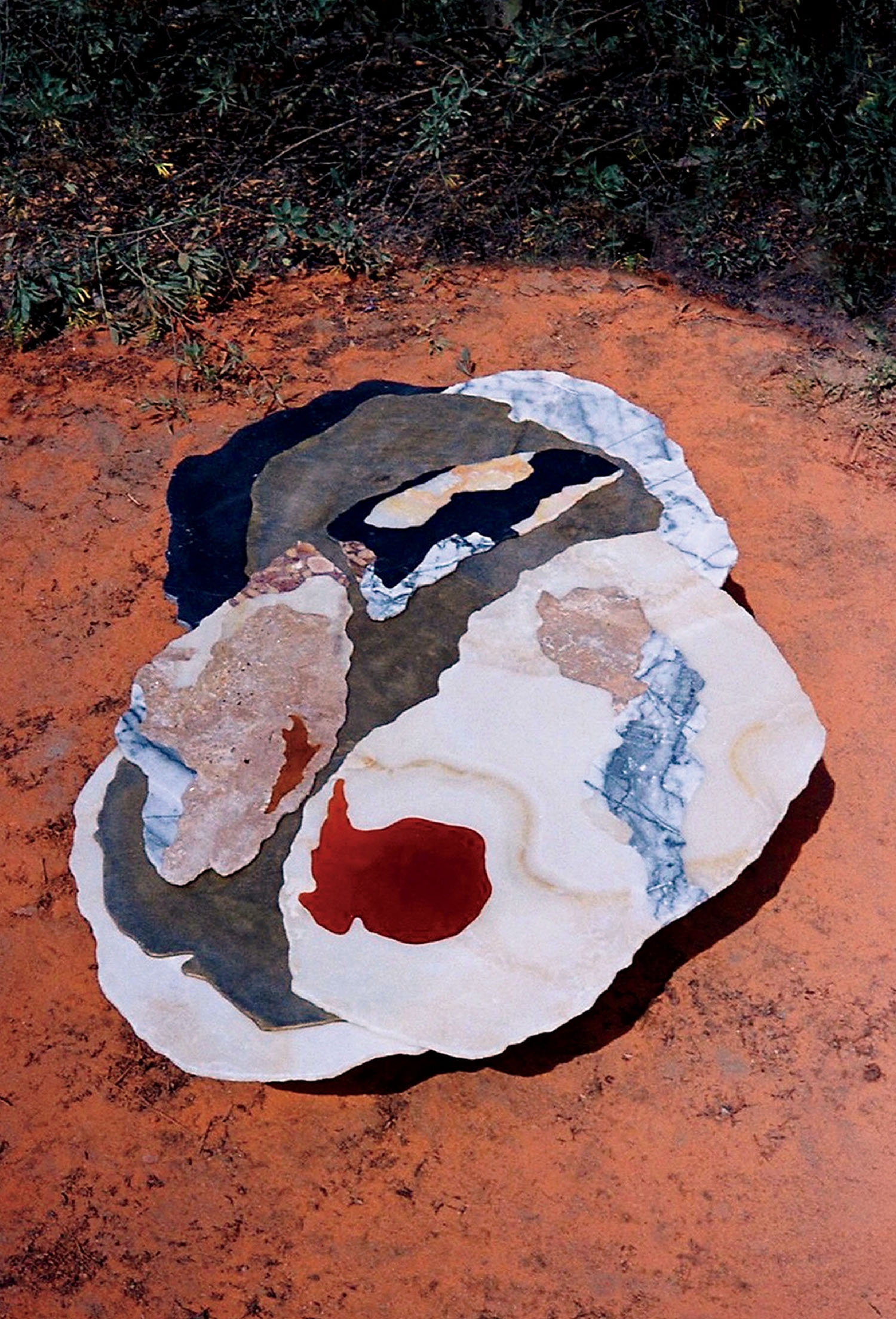 An abstract, multicolored stone sculpture with organic shapes sits on reddish ground, surrounded by sparse green vegetation.
