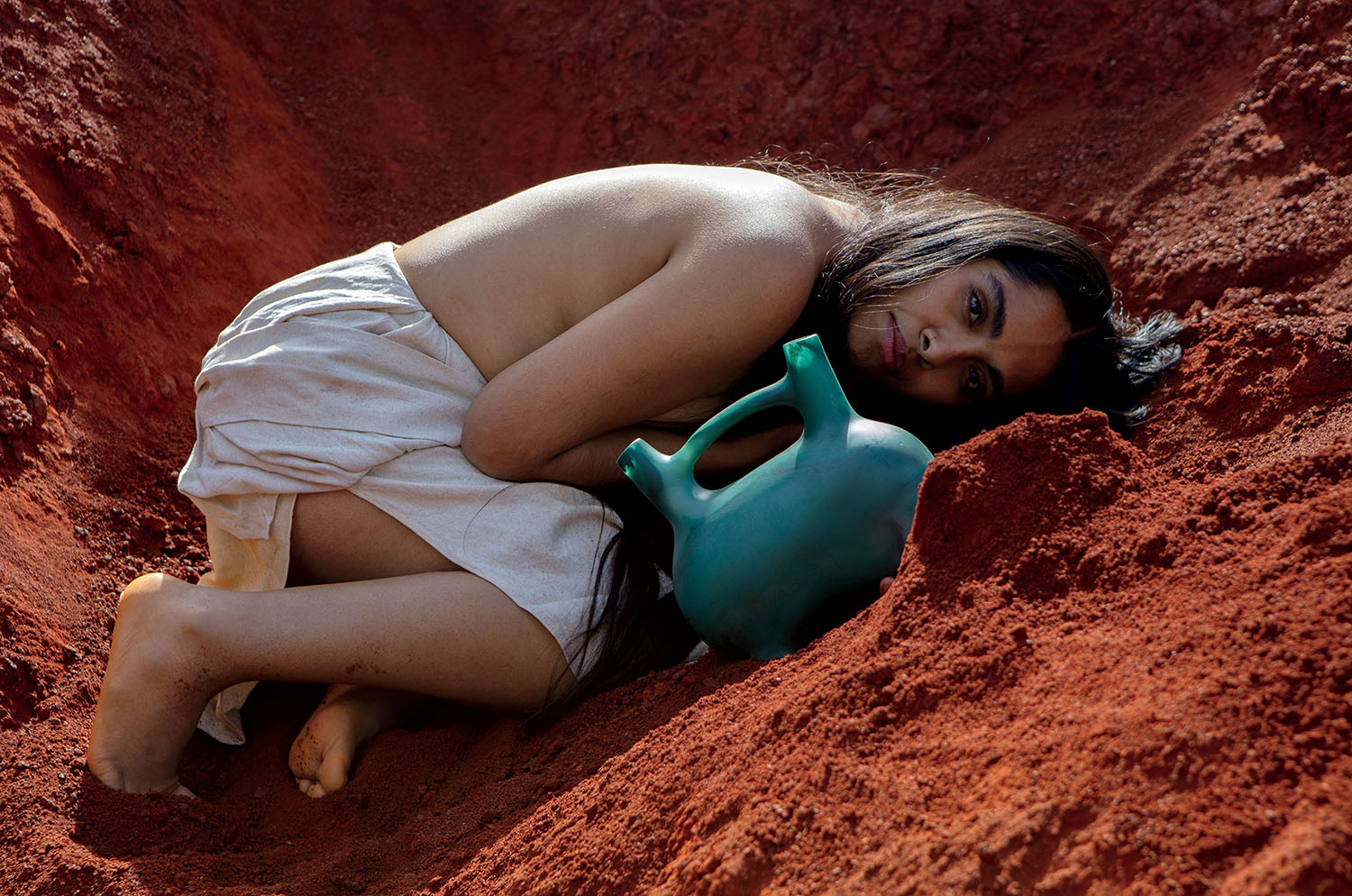 A person with long hair, draped in a white cloth, lies curled on red earth while holding a green ceramic vessel, looking directly at the camera.