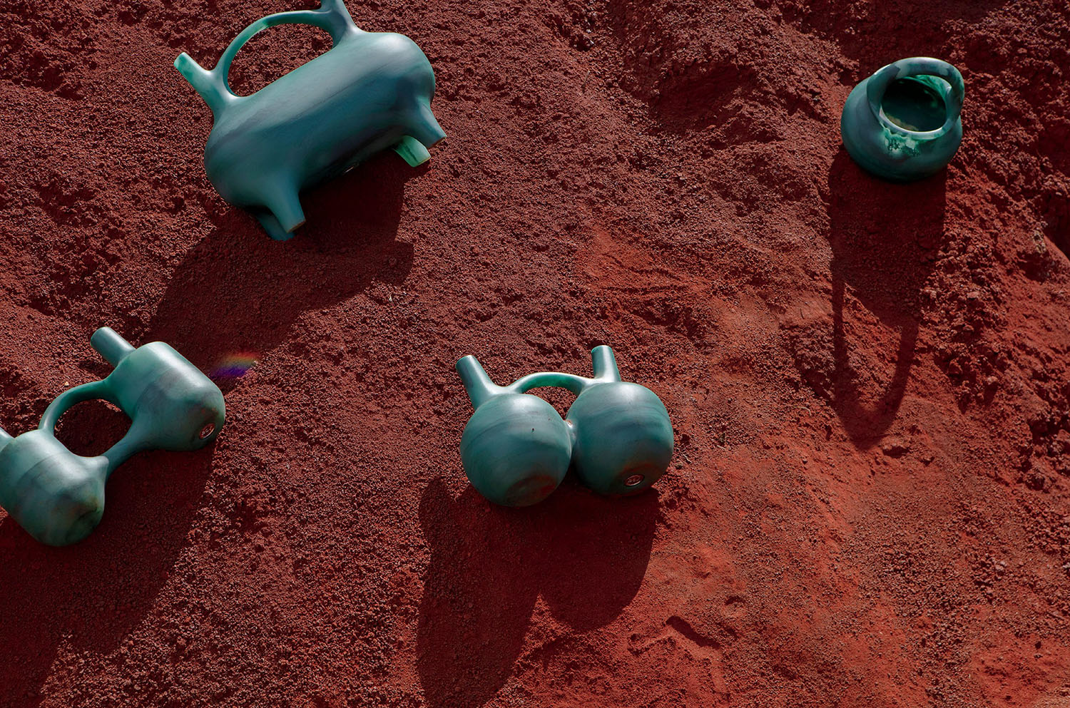 Four ceramic vessels with unique, abstract shapes are arranged on a textured surface of reddish-brown soil, casting distinct shadows.