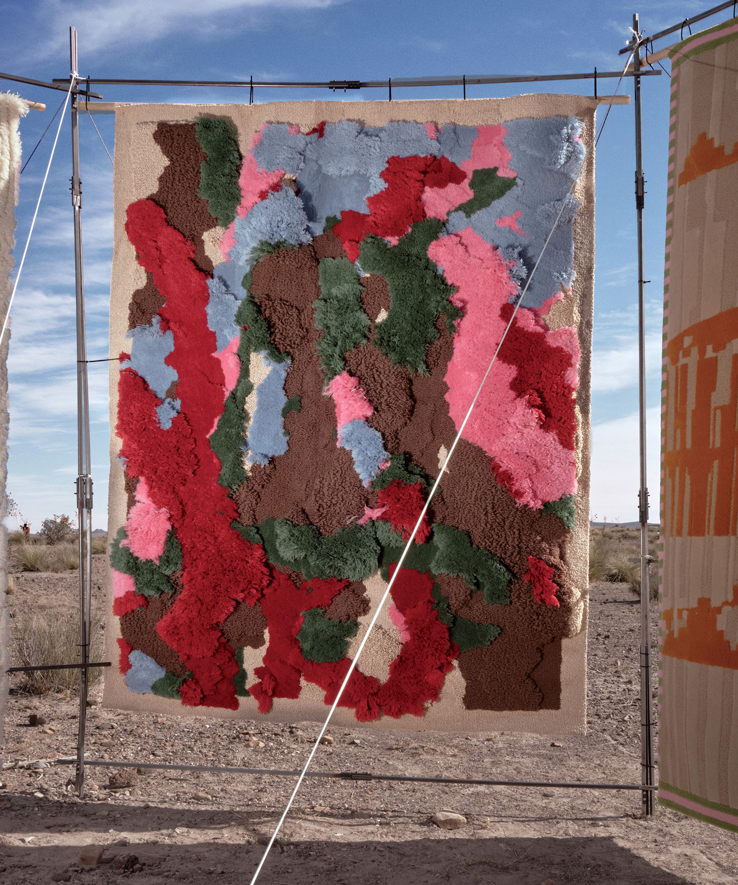 A textured, abstract rug with red, green, blue, pink, and brown patches hangs outdoors on a metal frame in a desert setting under a partly cloudy sky.