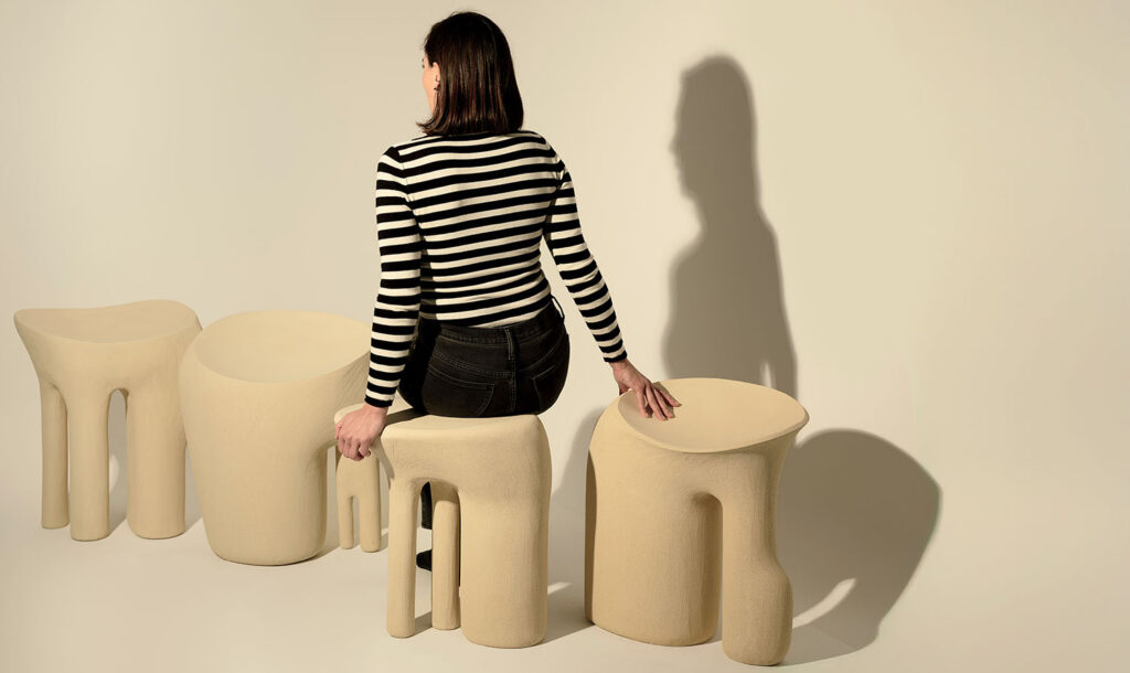 A woman in a black and white striped shirt sits on one of four beige, sculptural stools, facing away from the camera.