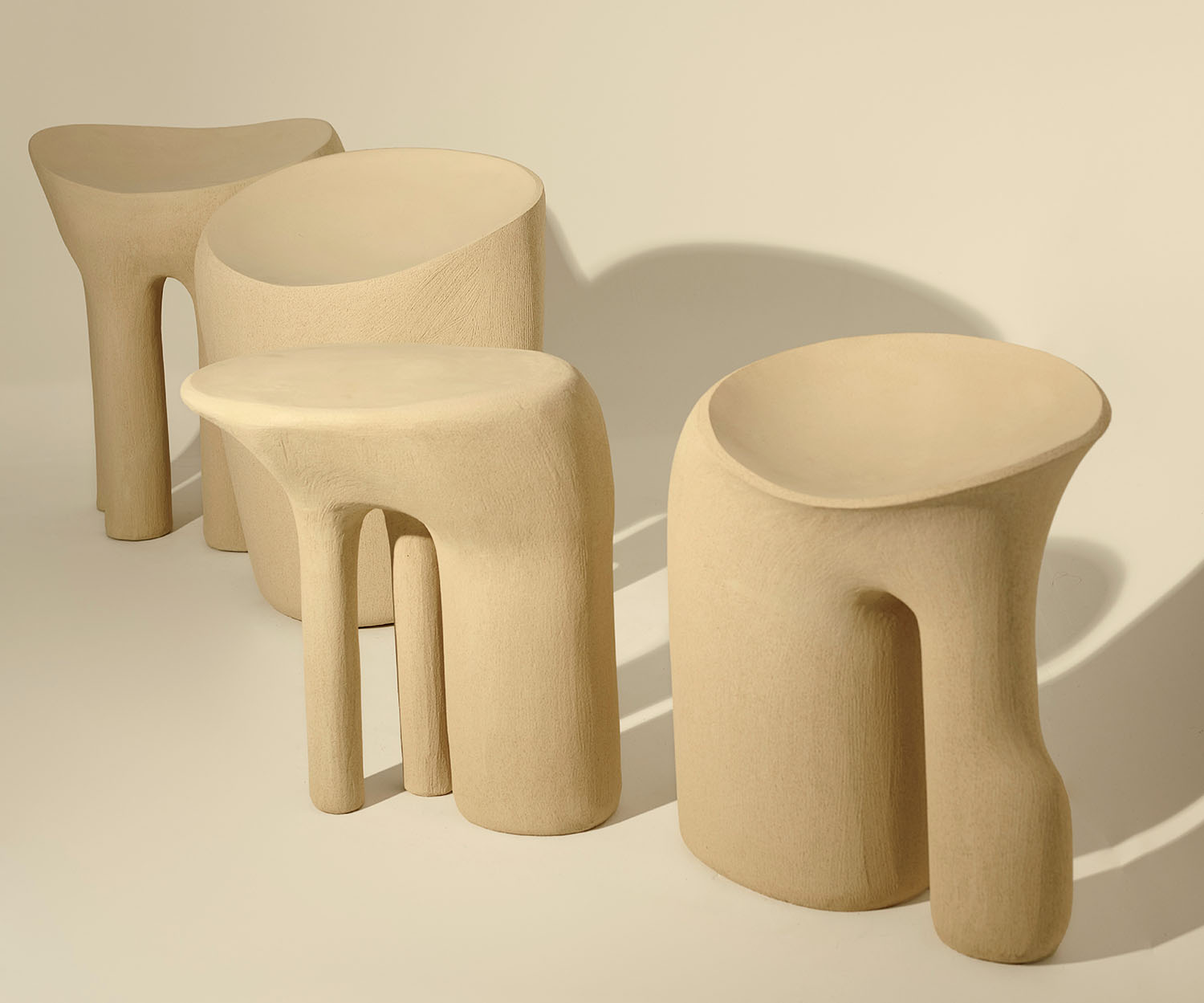 Four sculptural, beige stools with organic, asymmetrical shapes are arranged in a row on a neutral background.