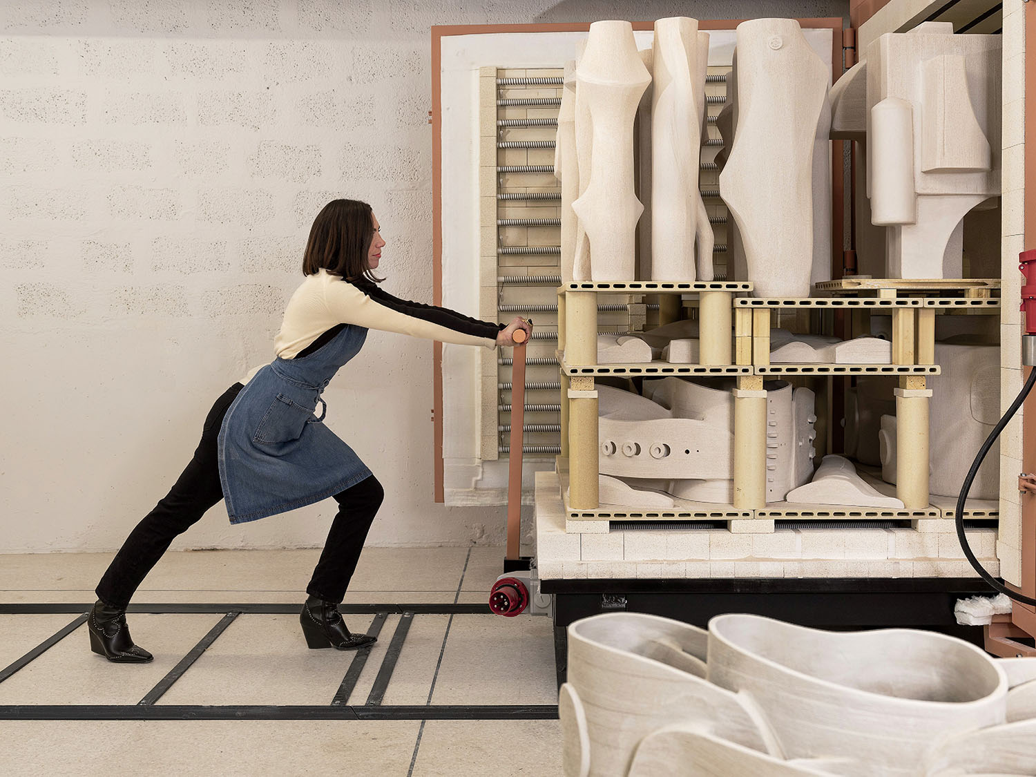 A woman in an apron pushes a cart loaded with large unglazed ceramic pieces into a kiln room.