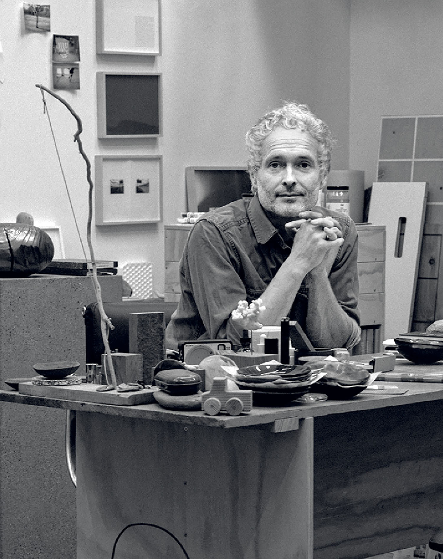 A man with curly hair and a beard sits at a cluttered desk in a studio, surrounded by art supplies and framed artworks on the wall behind him.
