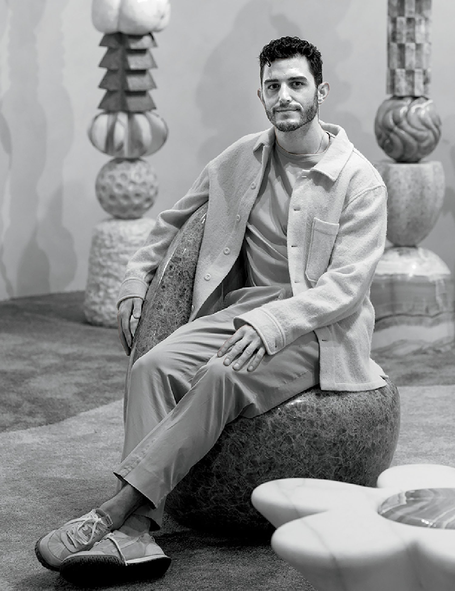 A man sits on a rounded stone-like seat in a room with abstract sculptures and textured objects, looking at the camera.