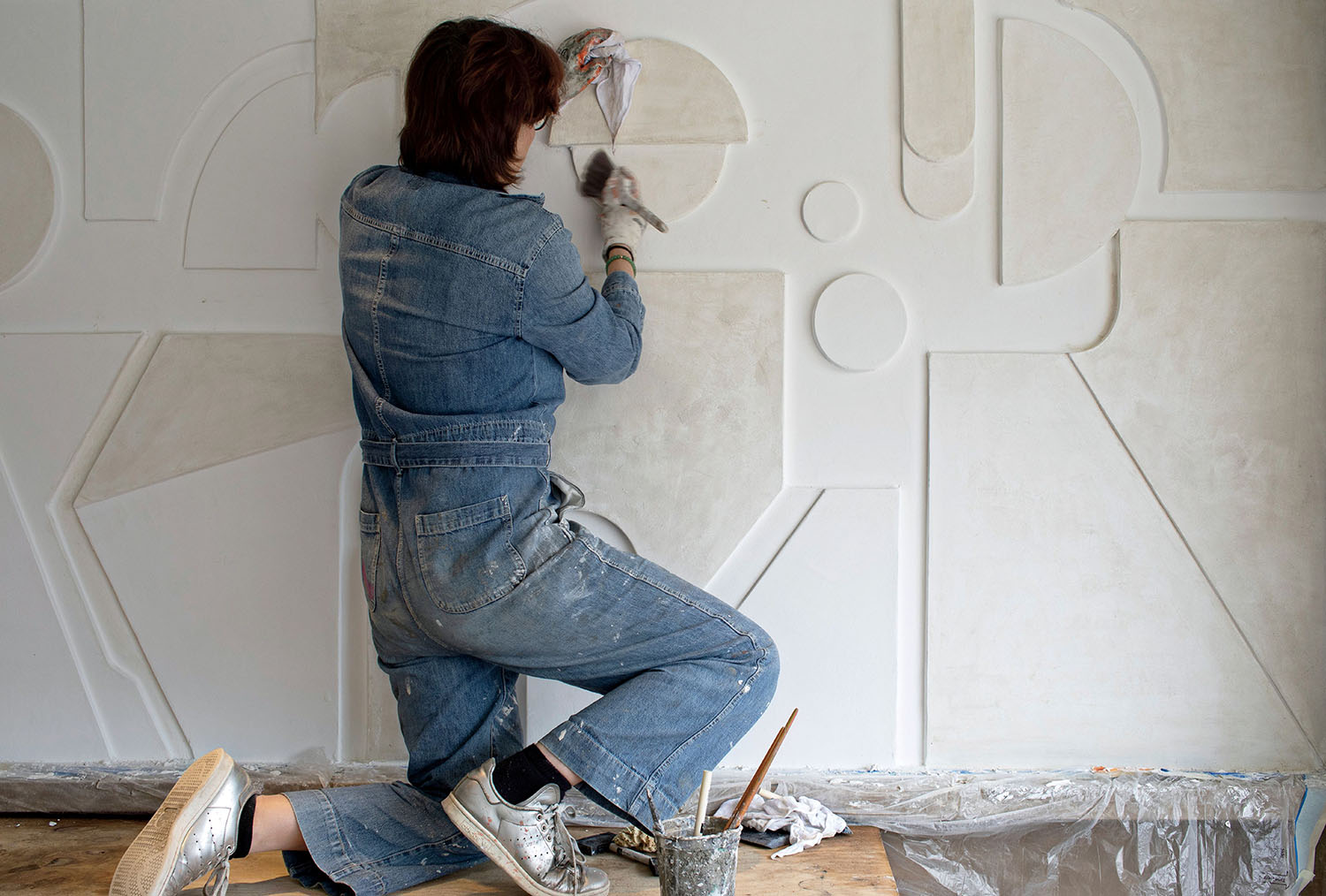 A person in denim overalls kneels while sculpting or painting geometric shapes on a large, textured white wall.
