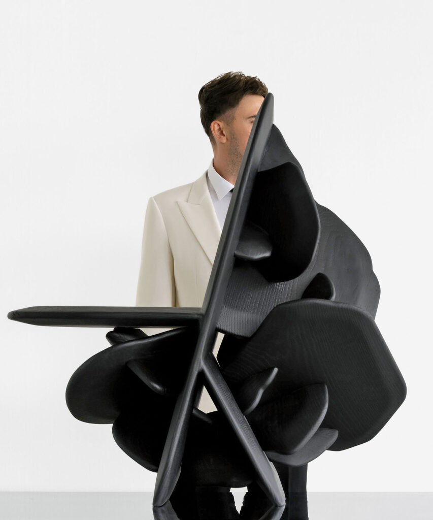 A man in a white suit stands behind a large abstract black sculpture, partially obscuring his face and body, against a plain white background.