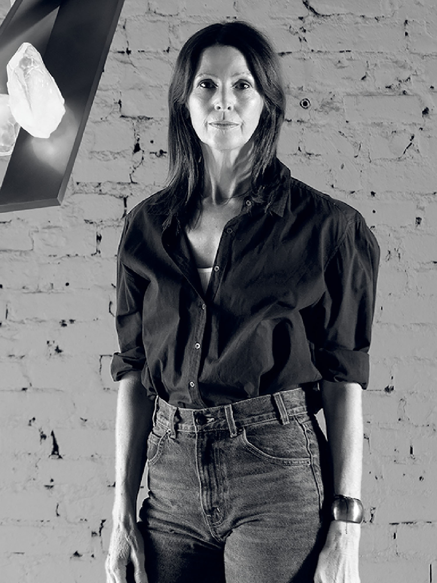 A woman with long dark hair stands in front of a brick wall, wearing a dark button-up shirt and jeans. A display with crystals appears on the left side.