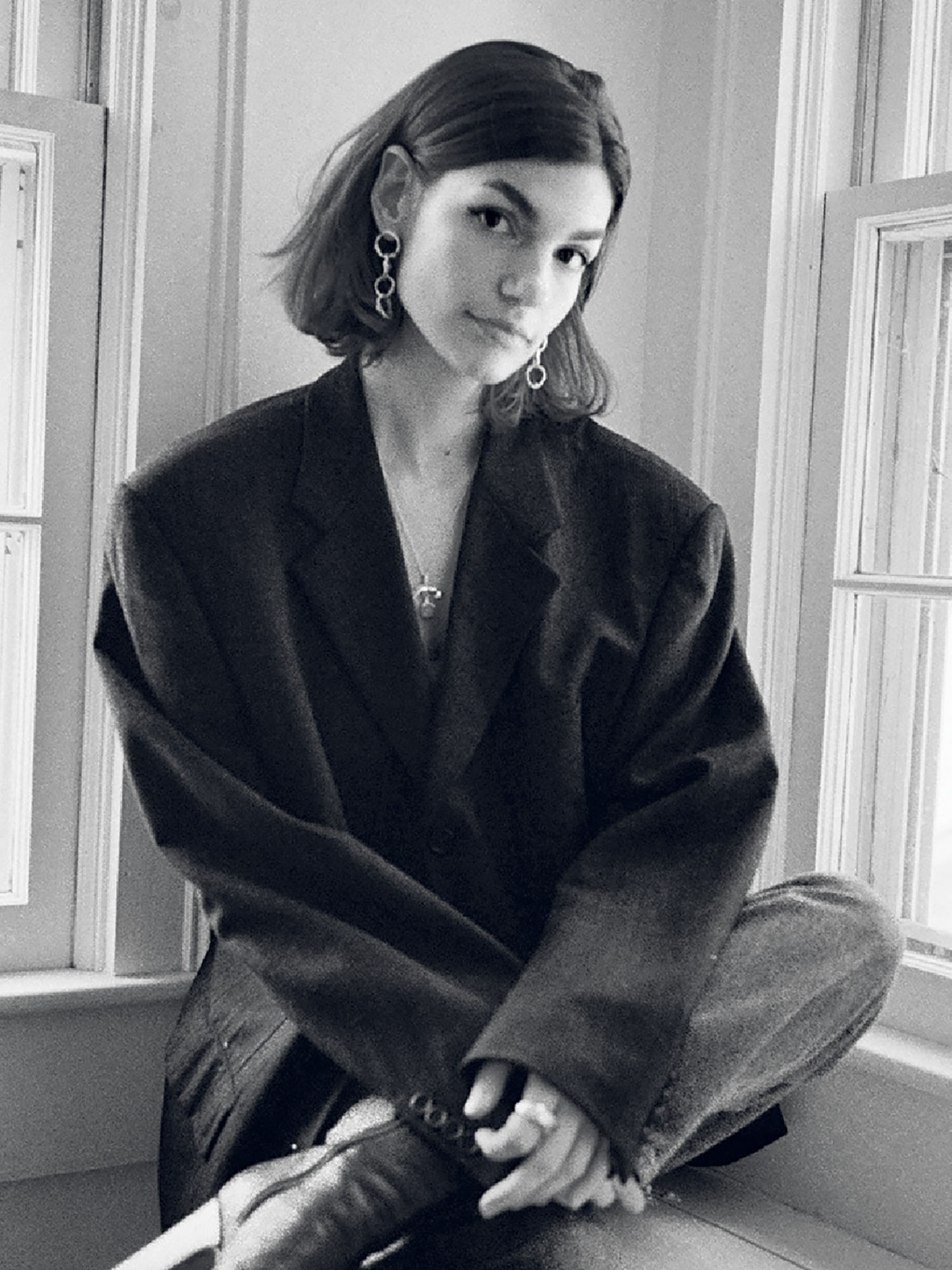 A person with short hair and earrings sits cross-legged on a windowsill, wearing an oversized blazer and jeans. The image is in black and white.