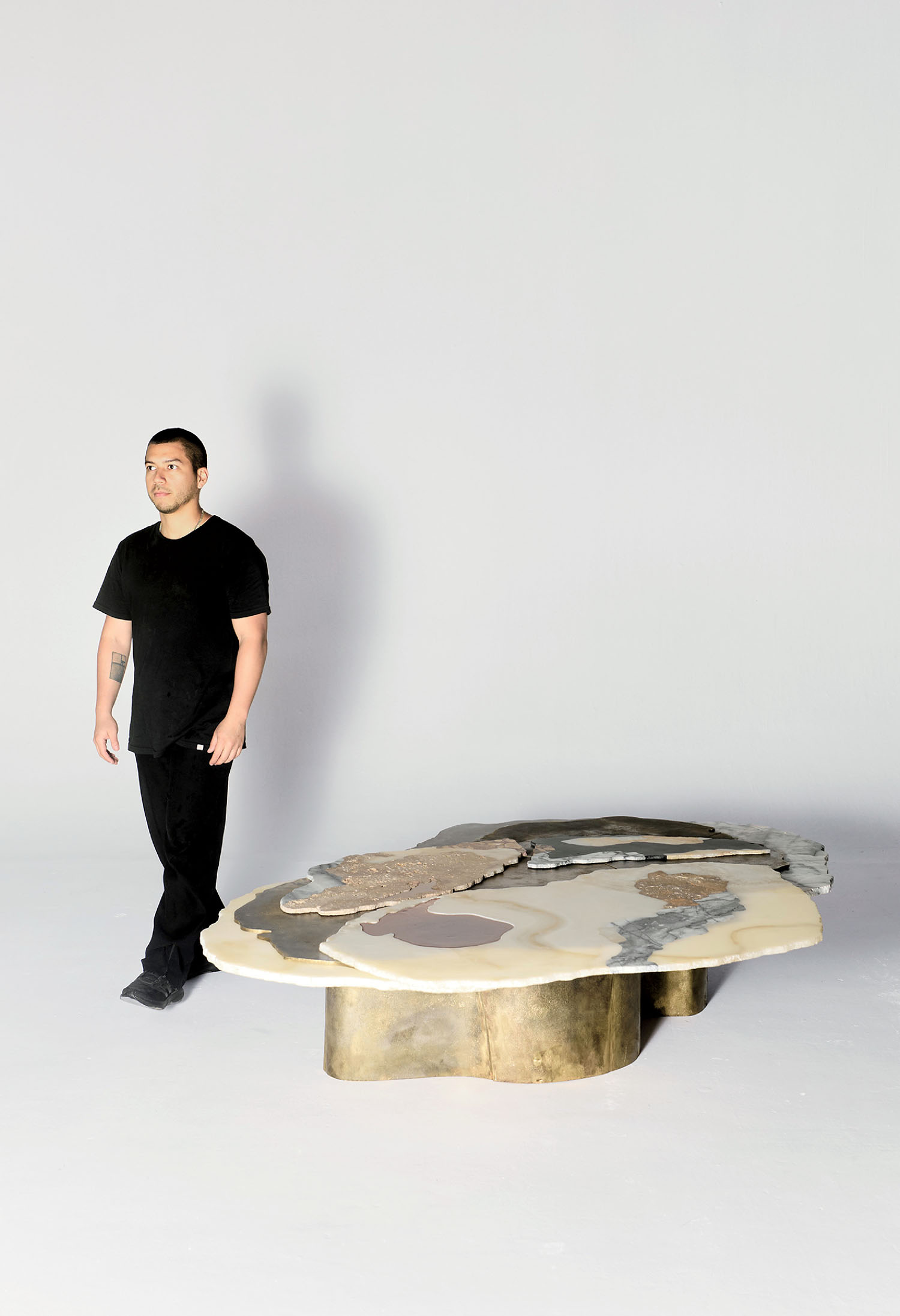 A person in black clothing stands next to a sculptural, multi-textured coffee table with a metallic base in a minimal white space.