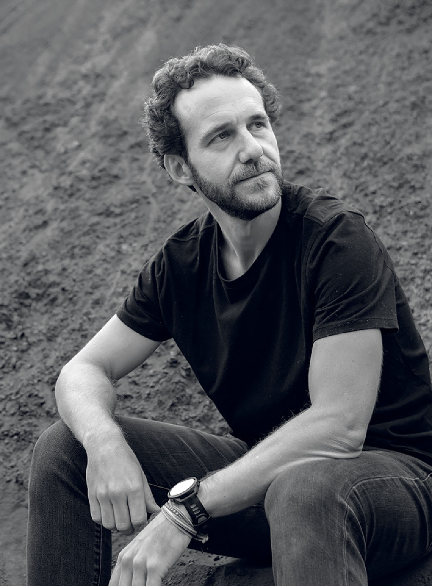 A man with curly hair and a beard, wearing a black t-shirt and jeans, sits outdoors on a slope, looking thoughtfully into the distance.