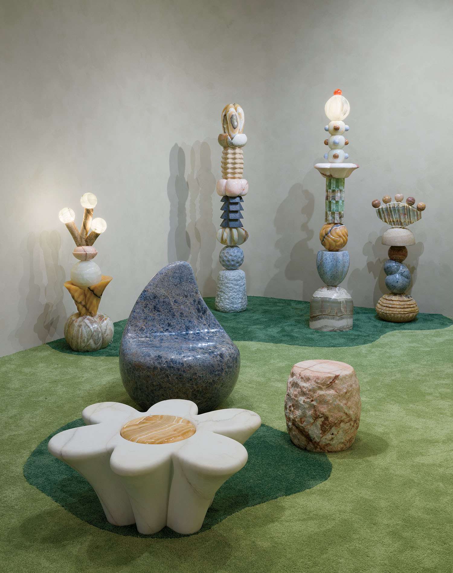 Colorful, abstract stone and marble sculptures and furniture are arranged on a green carpet in a gallery space with plain beige walls.