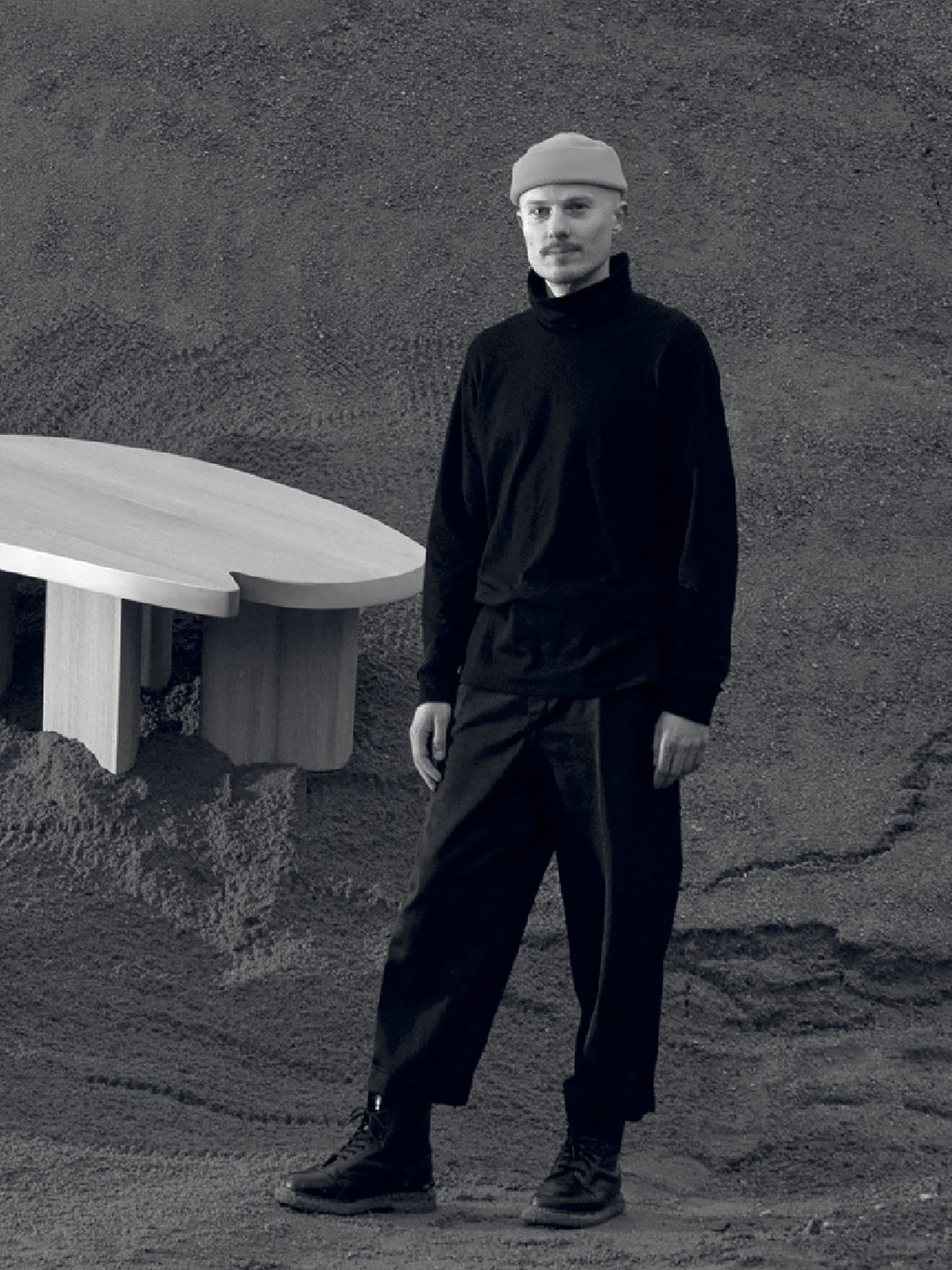 A person wearing a beanie, turtleneck, and loose pants stands next to a wooden table against a textured, earth-toned background.