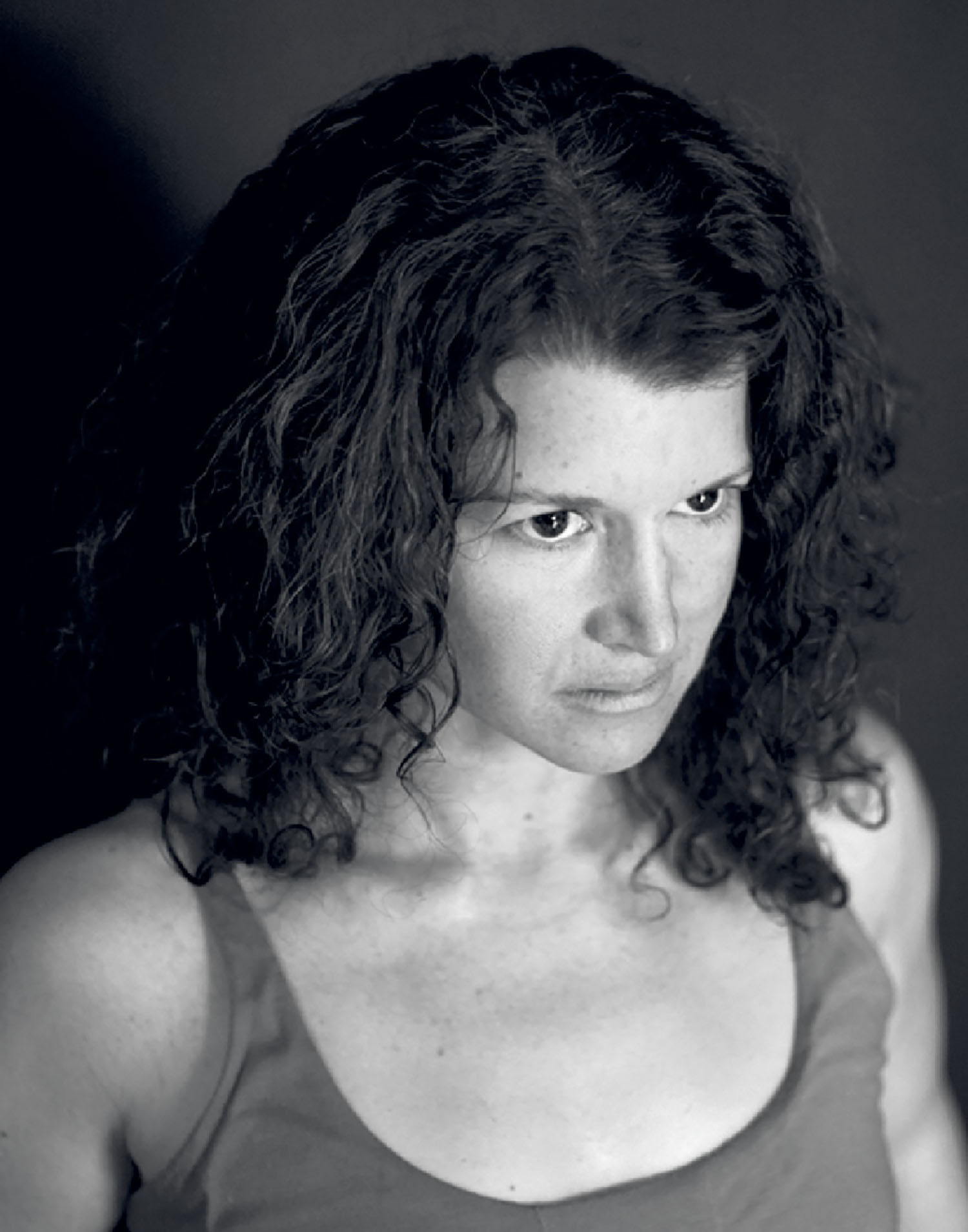 A woman with curly hair wearing a sleeveless top stares intently forward. The image is in black and white with a dark background.