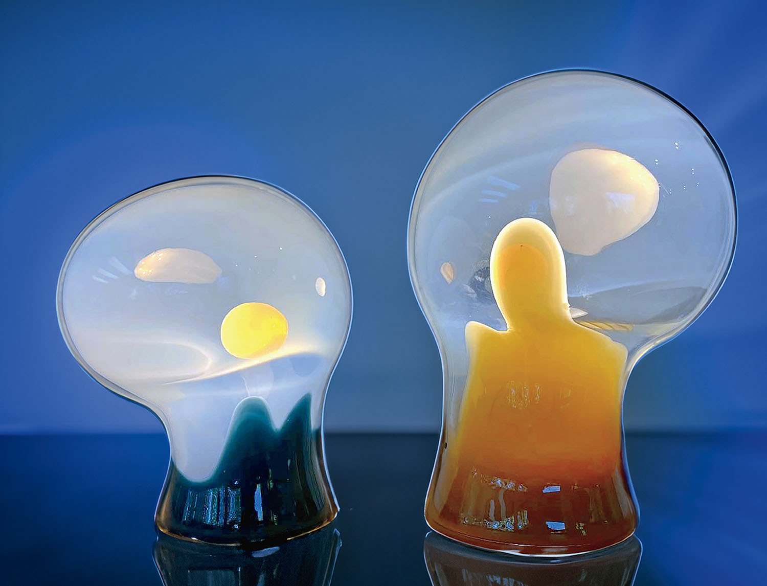 Two abstract glass sculptures with rounded shapes, featuring swirls of white, yellow, and orange, are displayed against a blue background.