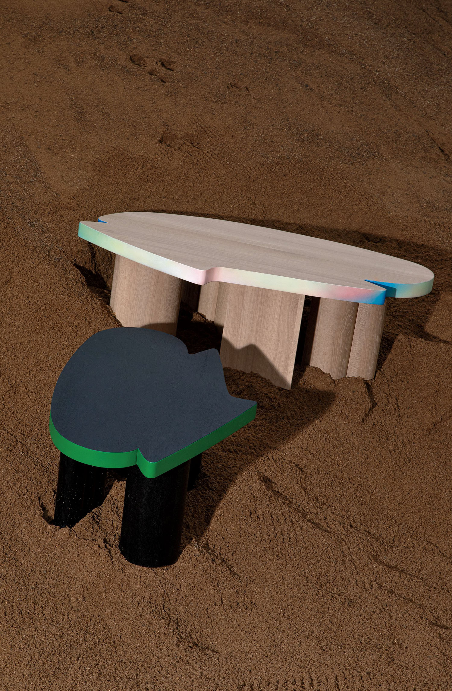 Two abstract, irregularly shaped wooden tables with painted edges are positioned on a sandy surface.