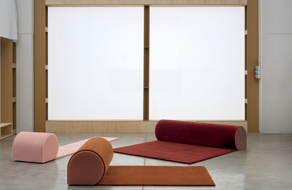 Three cylindrical cushions and matching rectangular mats in pink and burgundy are arranged on a concrete floor in front of large frosted windows.