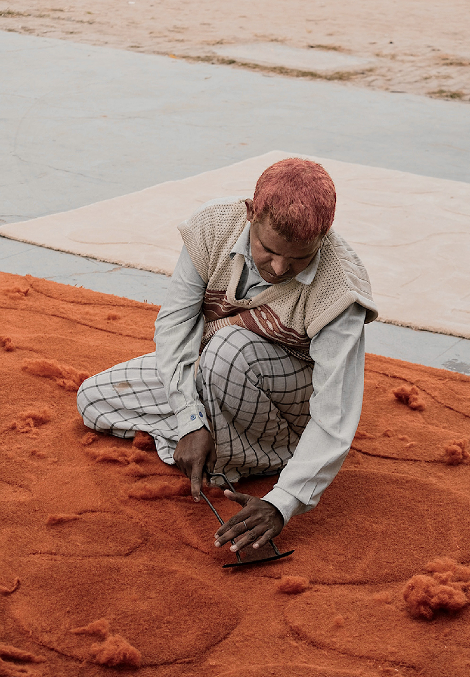 A person with dyed hair kneels on an orange carpet, using a metal tool to trim or shape its surface. Loose fibers are scattered around them.
