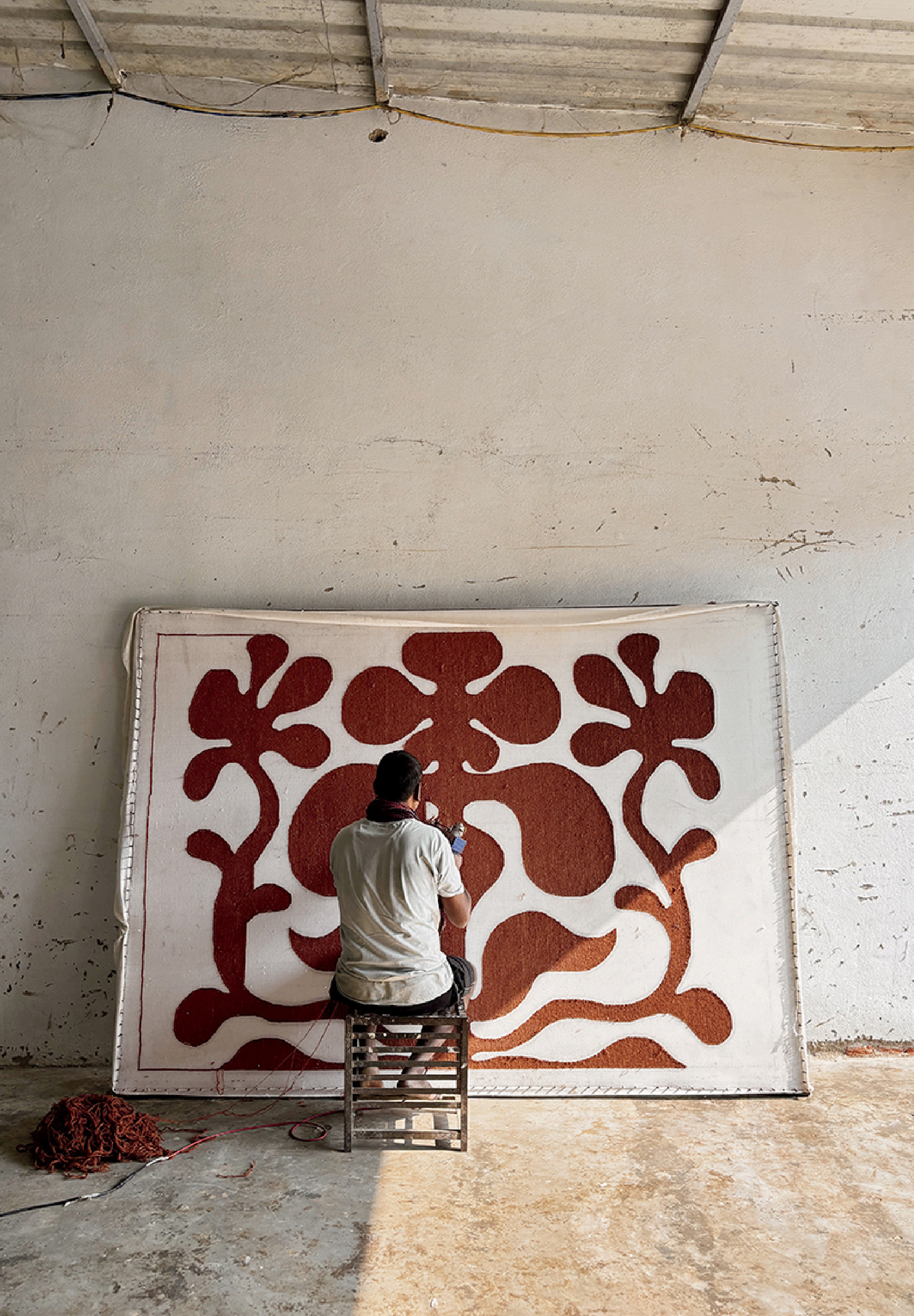 A person sits on a stool facing a large, abstract red and white textile artwork that leans against a bare wall in a spacious, light-filled room.