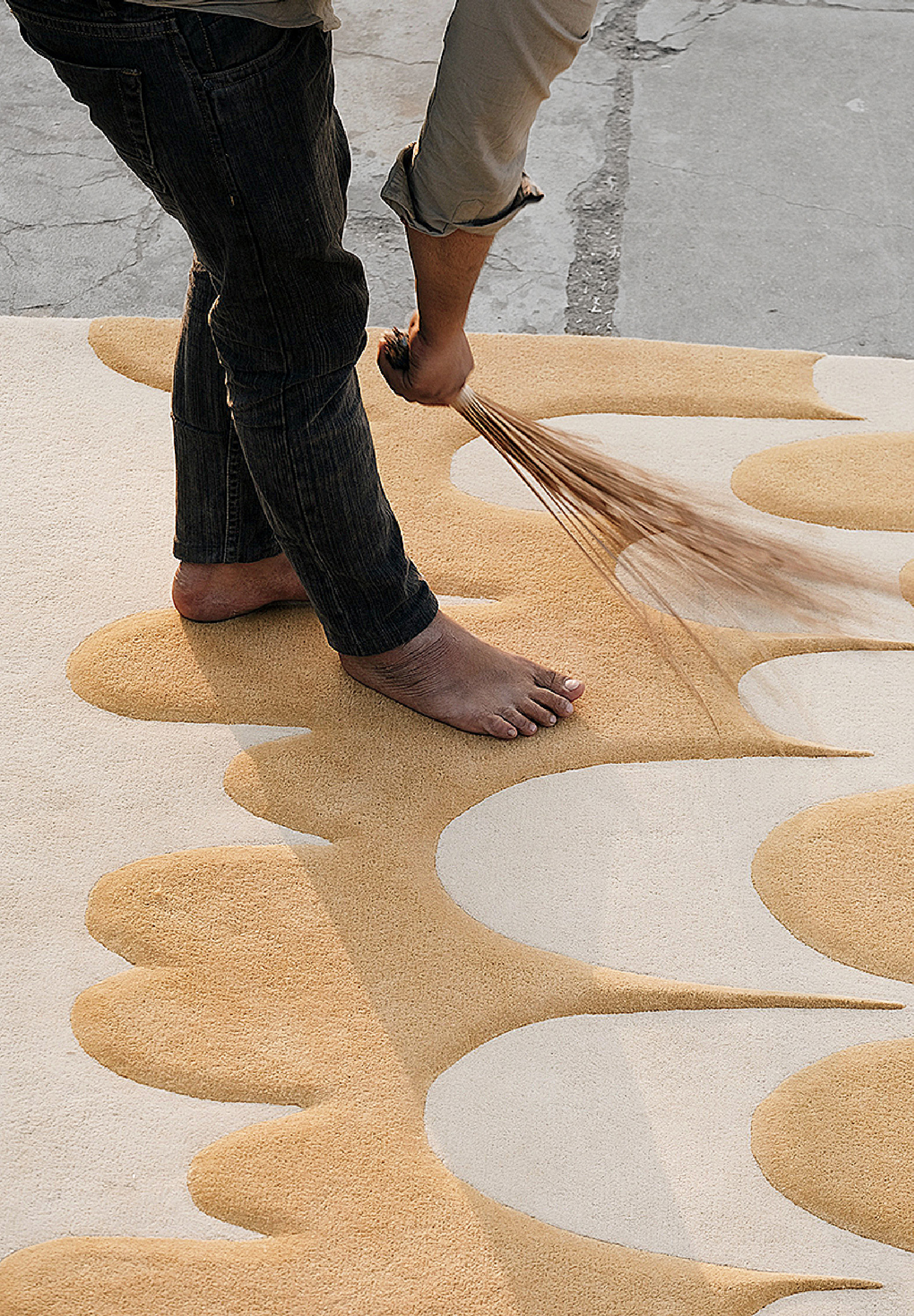 A person stands barefoot on a patterned rug while using a tool to brush or clean its surface.