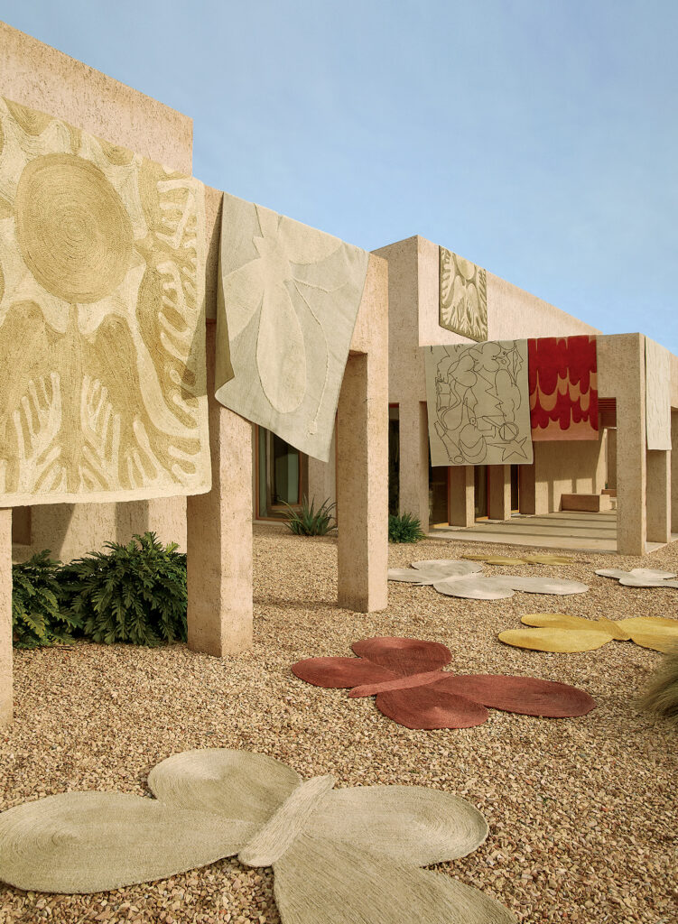 Large, textured rugs with abstract and botanical designs hang from a beige stone building, with more rugs in various shapes laid out on the gravel ground.
