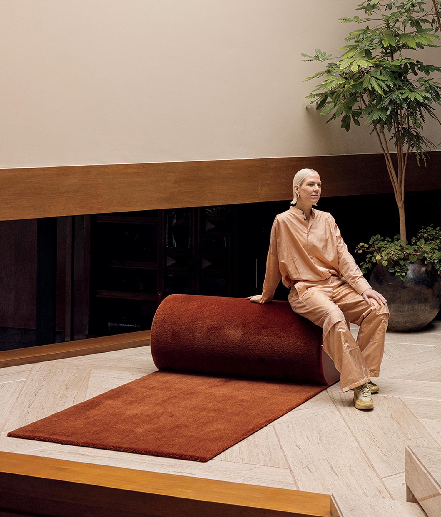 A person in peach-colored clothing sits on a cylindrical red rug in a modern interior with beige walls, wooden accents, and a potted plant.