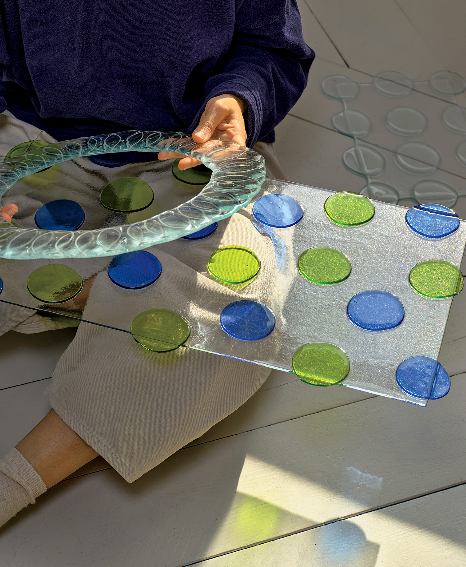 A person seated on the floor holds a round glass plate and a rectangular glass sheet with blue and green circular patterns.