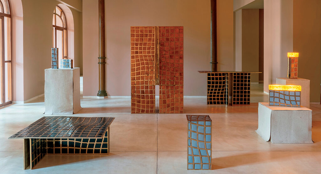 Art gallery interior featuring modern sculptures and furniture with geometric, grid-like patterns in earthy tones, displayed on concrete plinths and a minimalist floor.