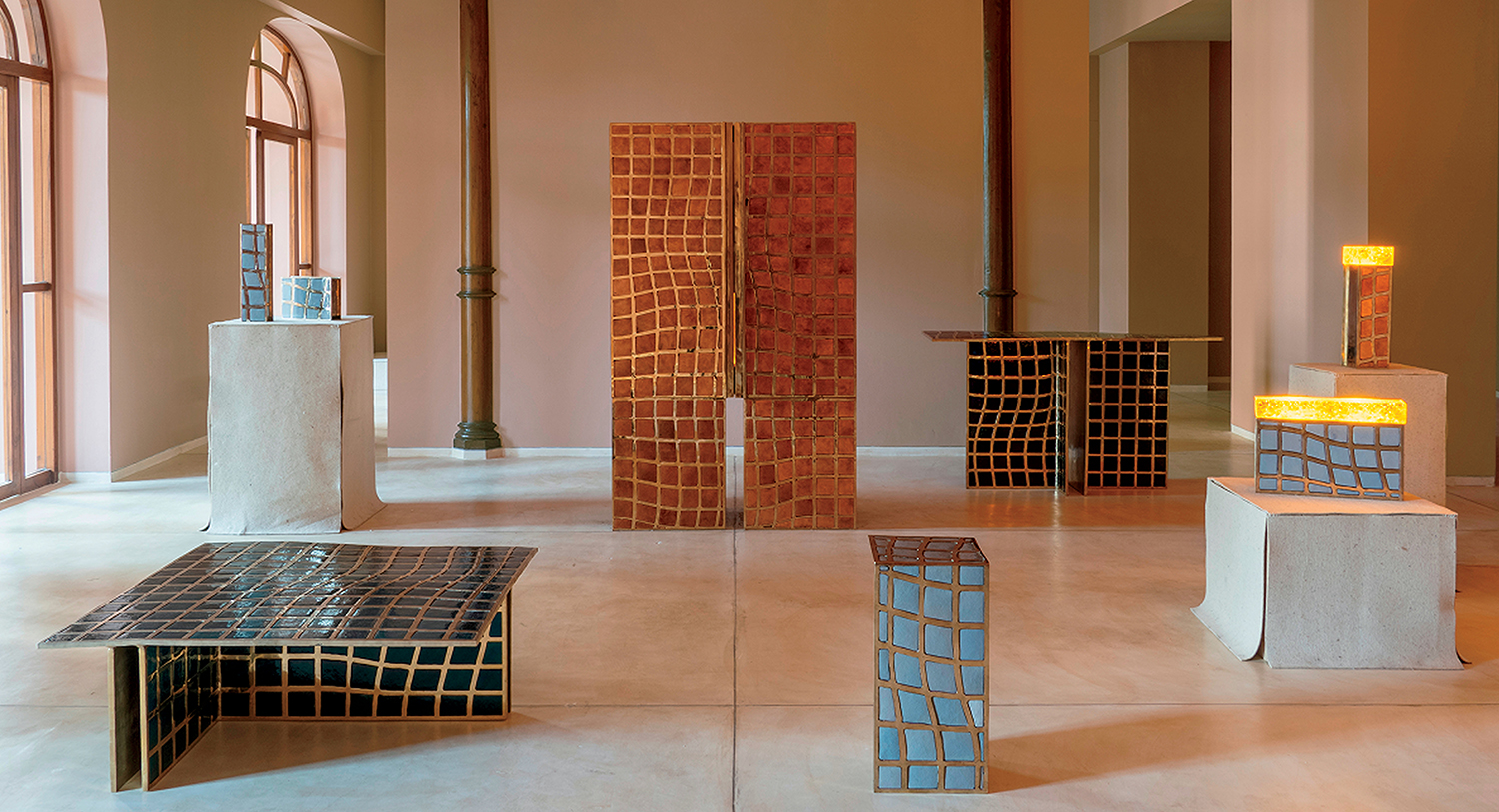 Art gallery interior featuring modern sculptures and furniture with geometric, grid-like patterns in earthy tones, displayed on concrete plinths and a minimalist floor.